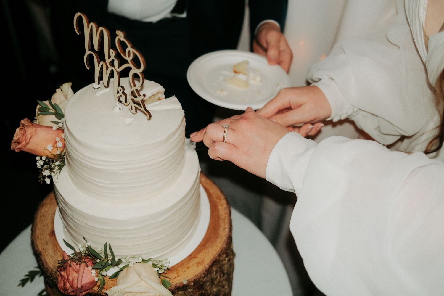 A white tiered wedding cake with Mr and Mrs topper being cut on a rustic wood slice with pink floral accents.