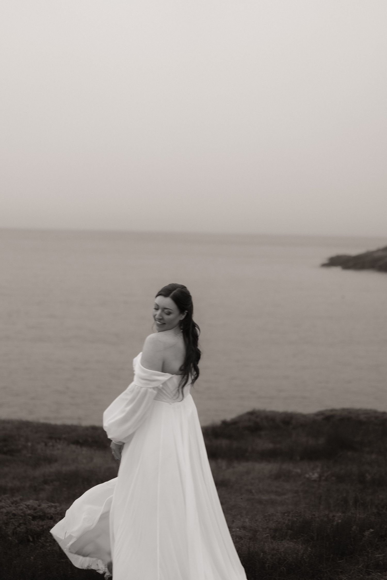 A flowing white dress billows in the wind against a moody coastal landscape in black and white.