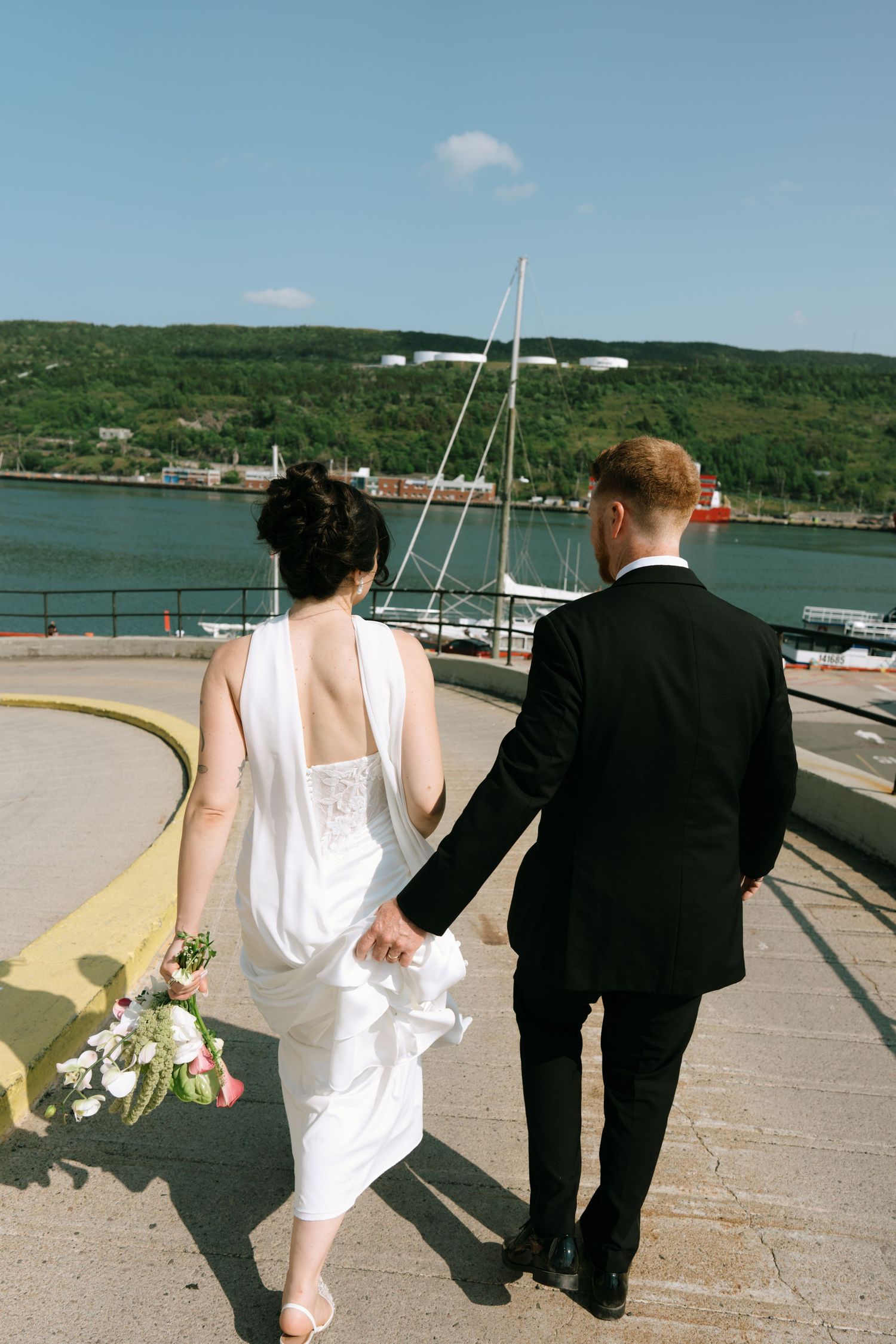 Bride and groom walking hand in hand on a waterfront pier, with a sailboat and green hills in the background.