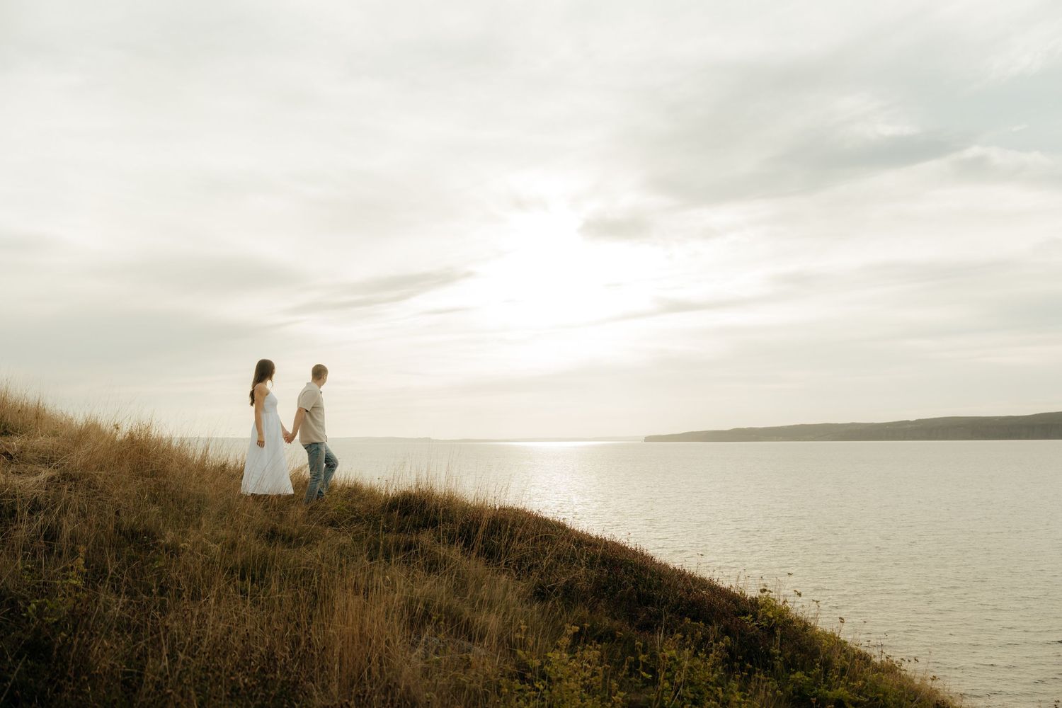 Two silhouettes stand on a grassy hillside overlooking a serene lake at sunset.