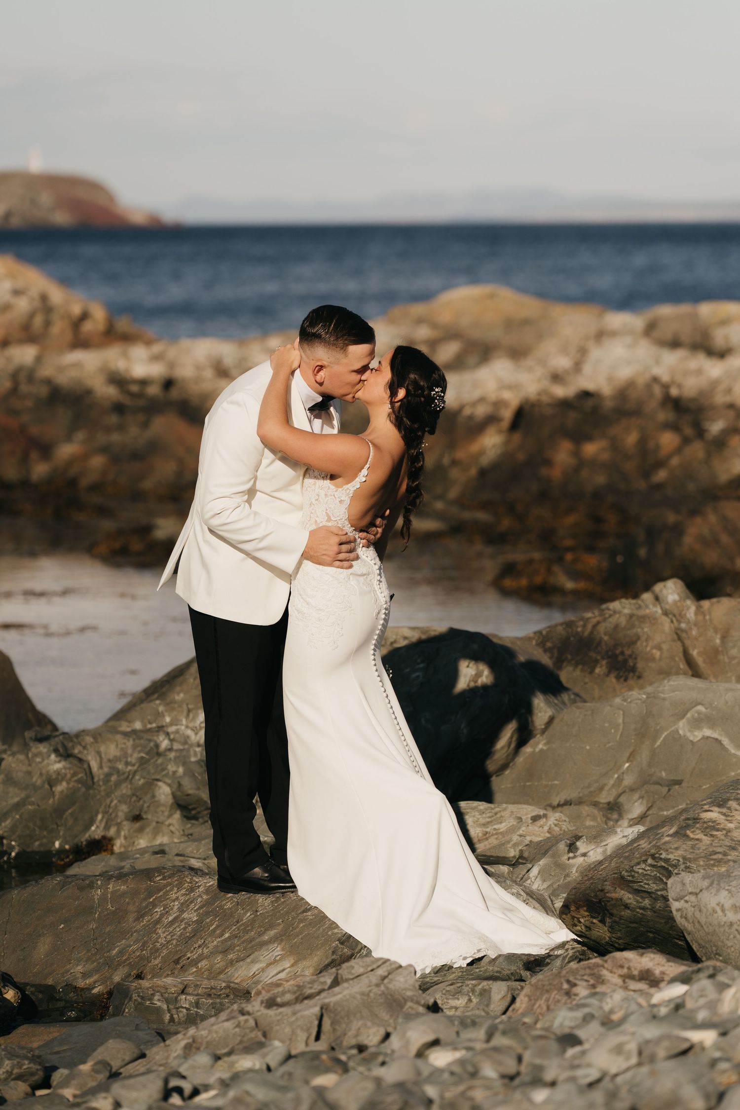 A romantic wedding couple shares intimate moments on a rocky beach coastline at sunset.