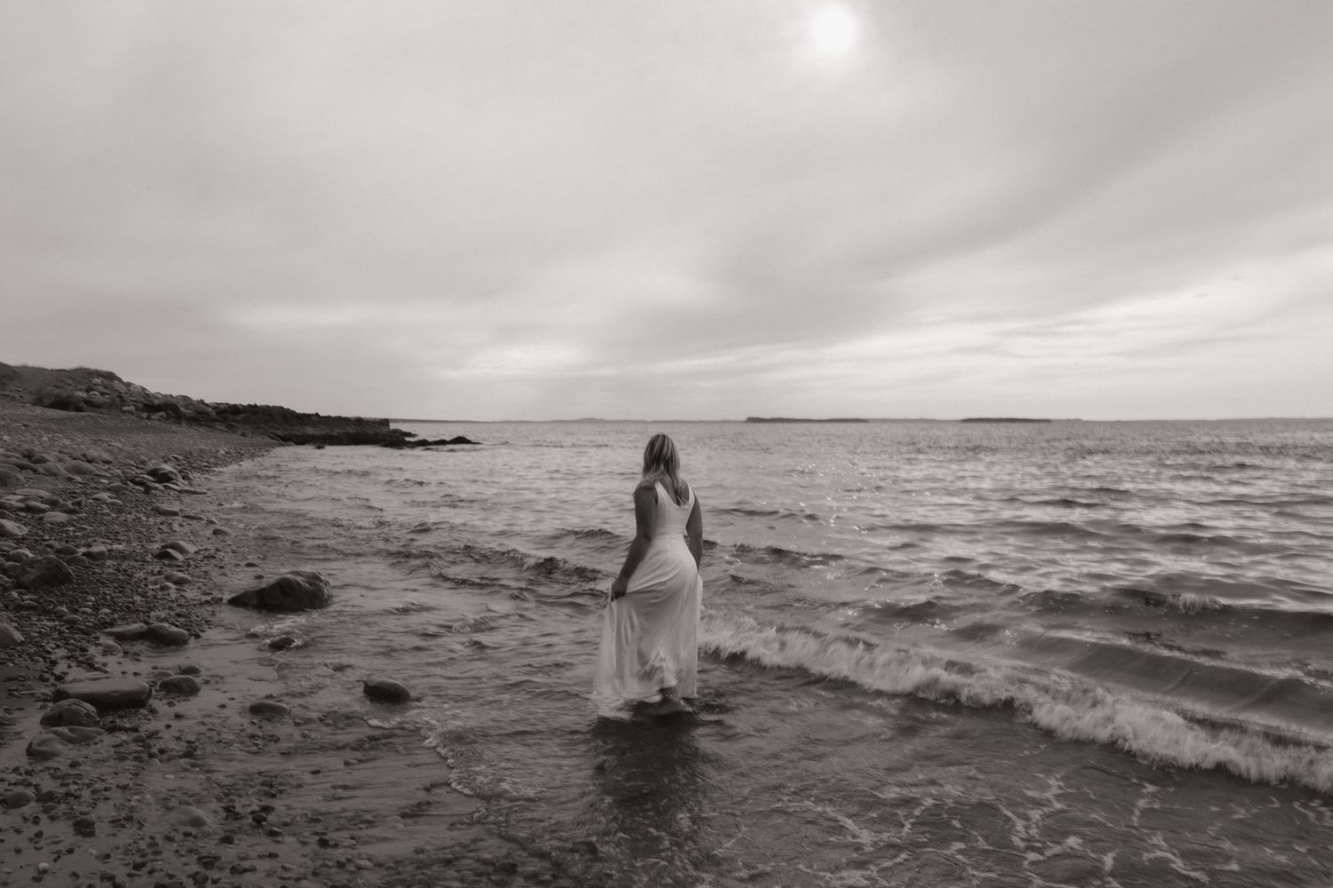 A bride in a white dress stands alone on a rocky beach shore during an overcast day.