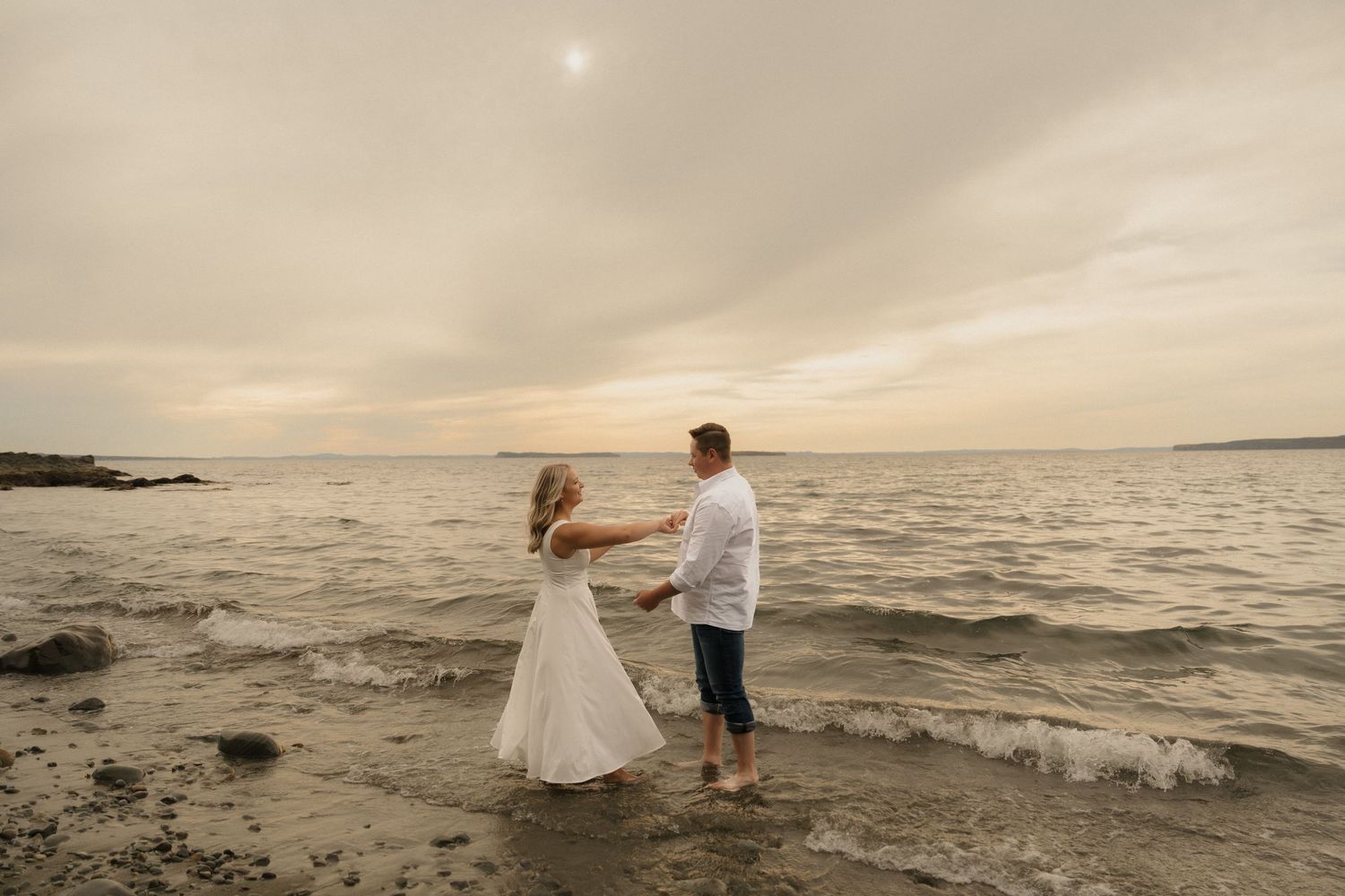 Romantic couple stands in shallow ocean water during sunset with waves crashing around them.