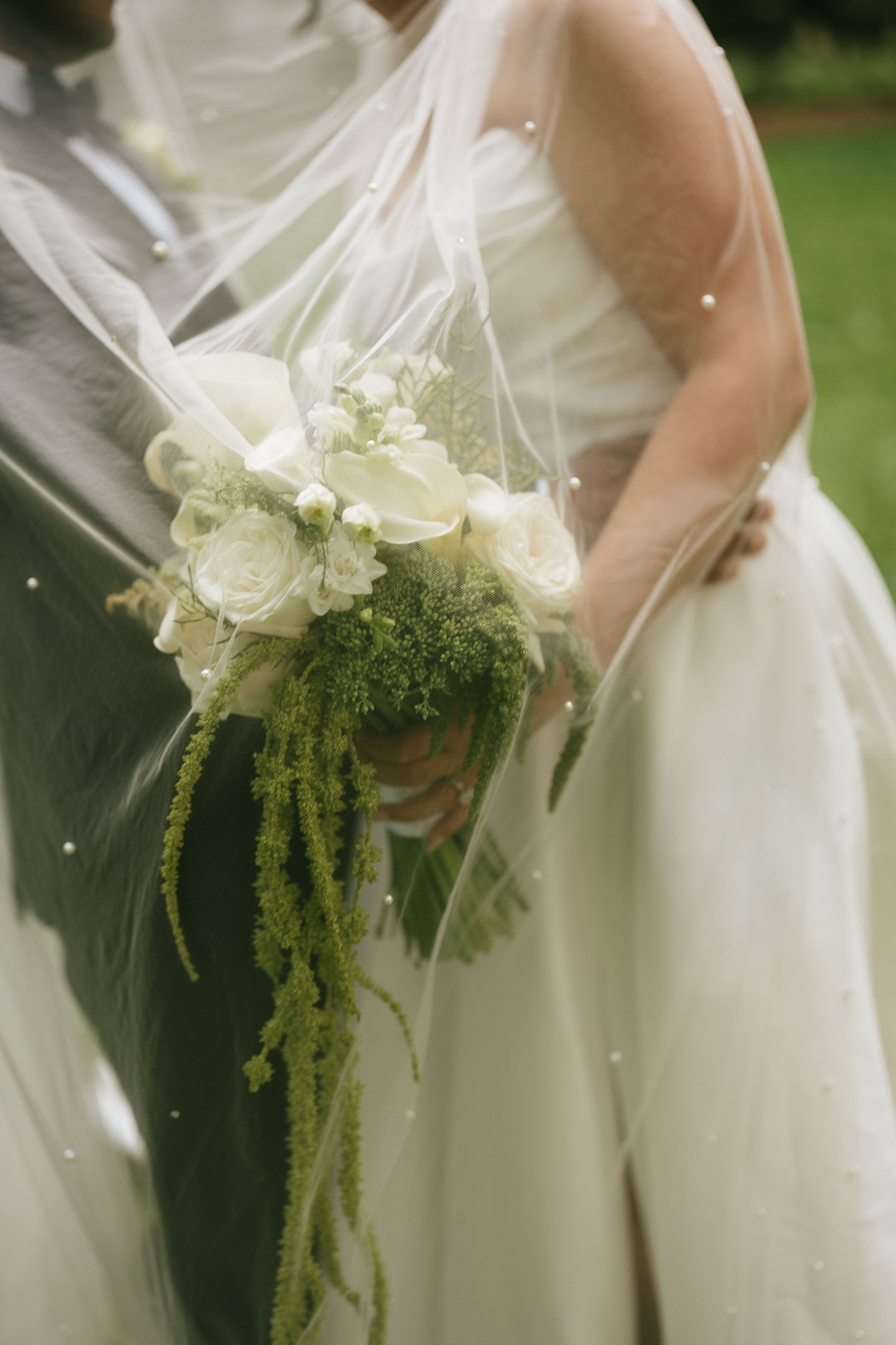 Bride holding elegant white rose and green cascading wedding bouquet, draped with sheer veil, standing beside groom outdoors.