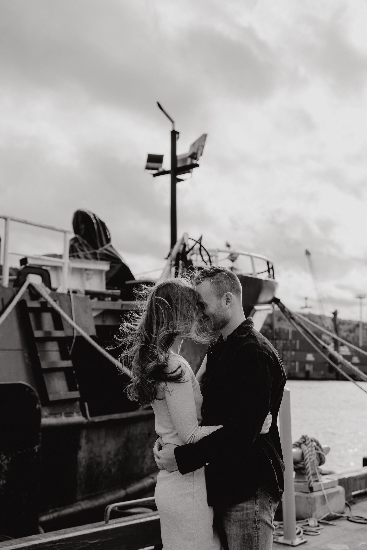 Black and white portrait of couple sharing an intimate moment by harbor ships.