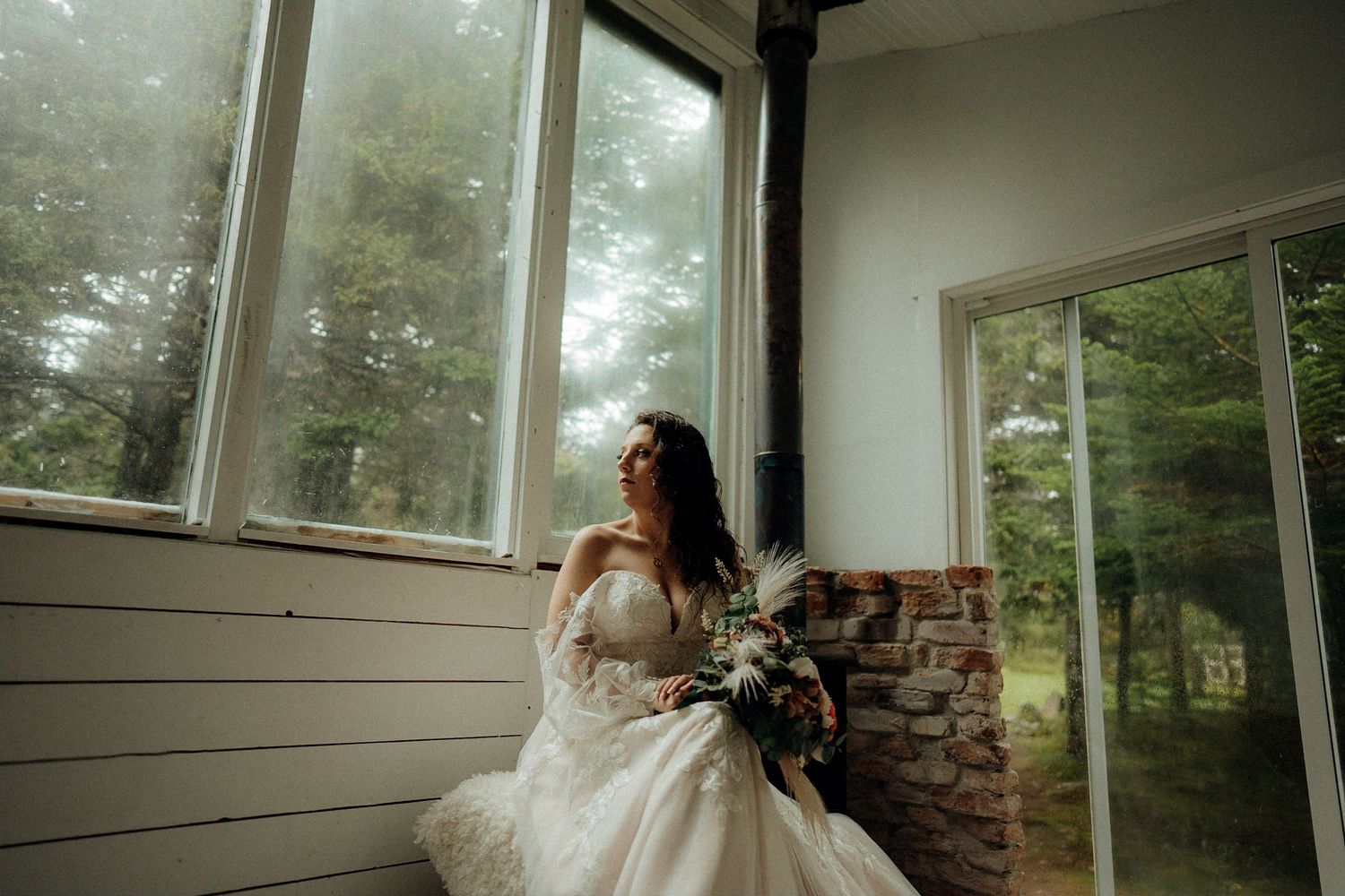 Bride sits in sunlit window seat holding elegant floral bouquet.