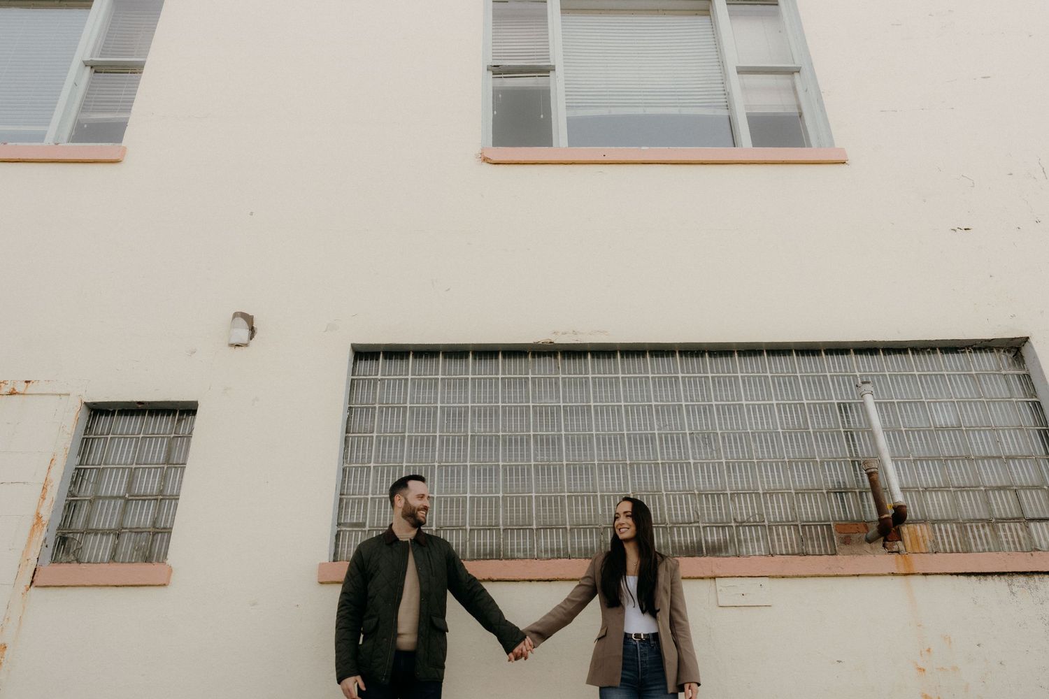 Couple holding hands and smiling at each other in front of a white building with large windows.