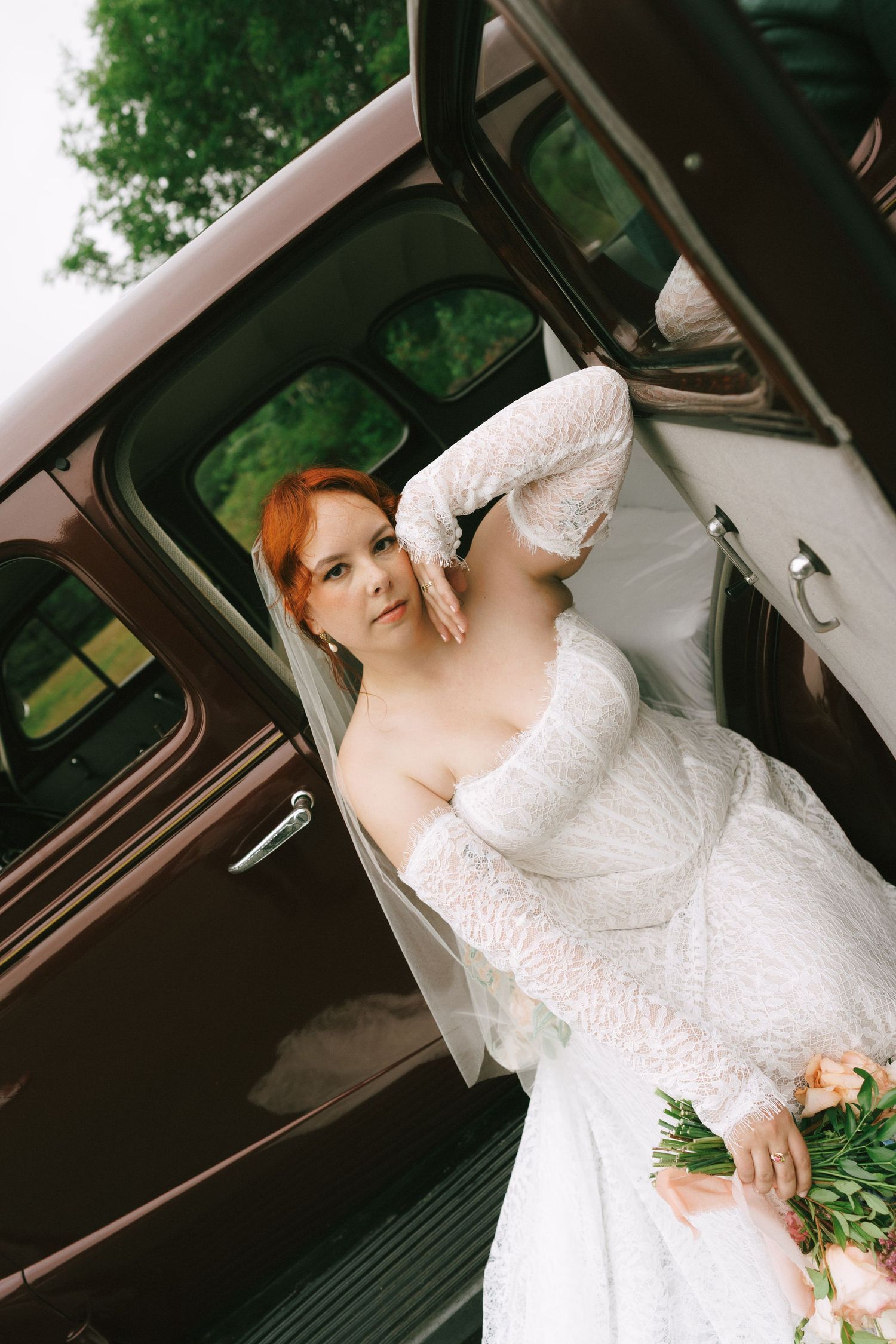 Red-haired bride in white lace gown holding bouquet, posing beside vintage brown car door outdoors.