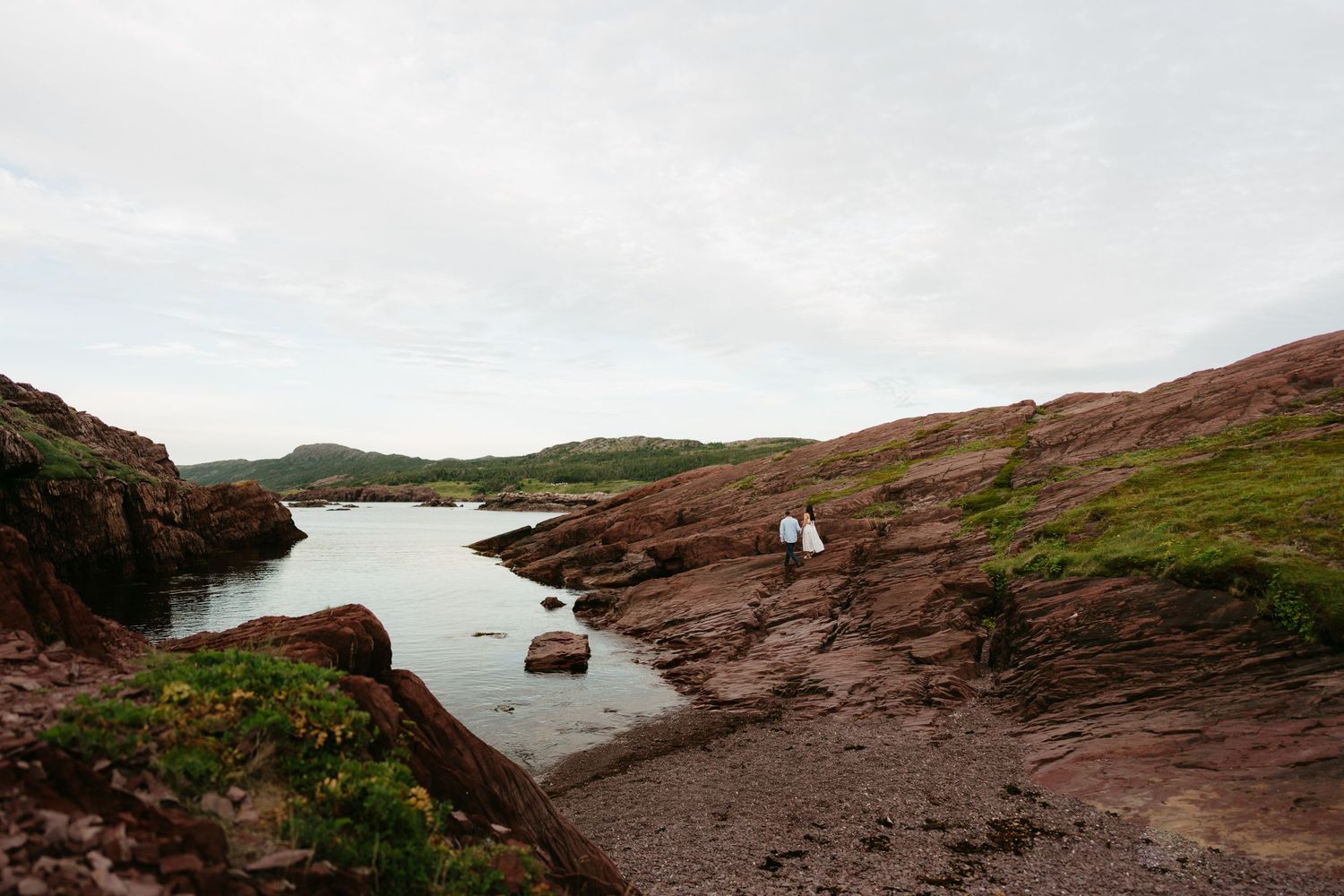 Couple standing on red rocky coastal cliffs beside calm water with green hills in the background.