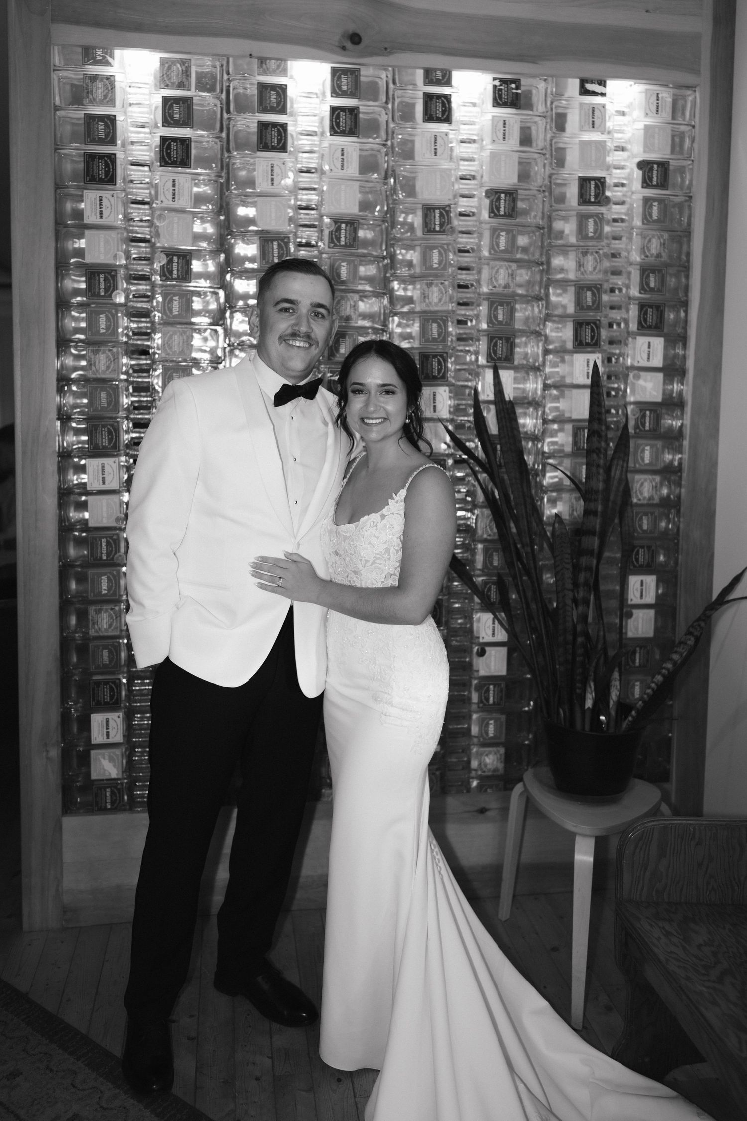 Black and white wedding photo showing couple embracing in front of wall covered with framed photographs.