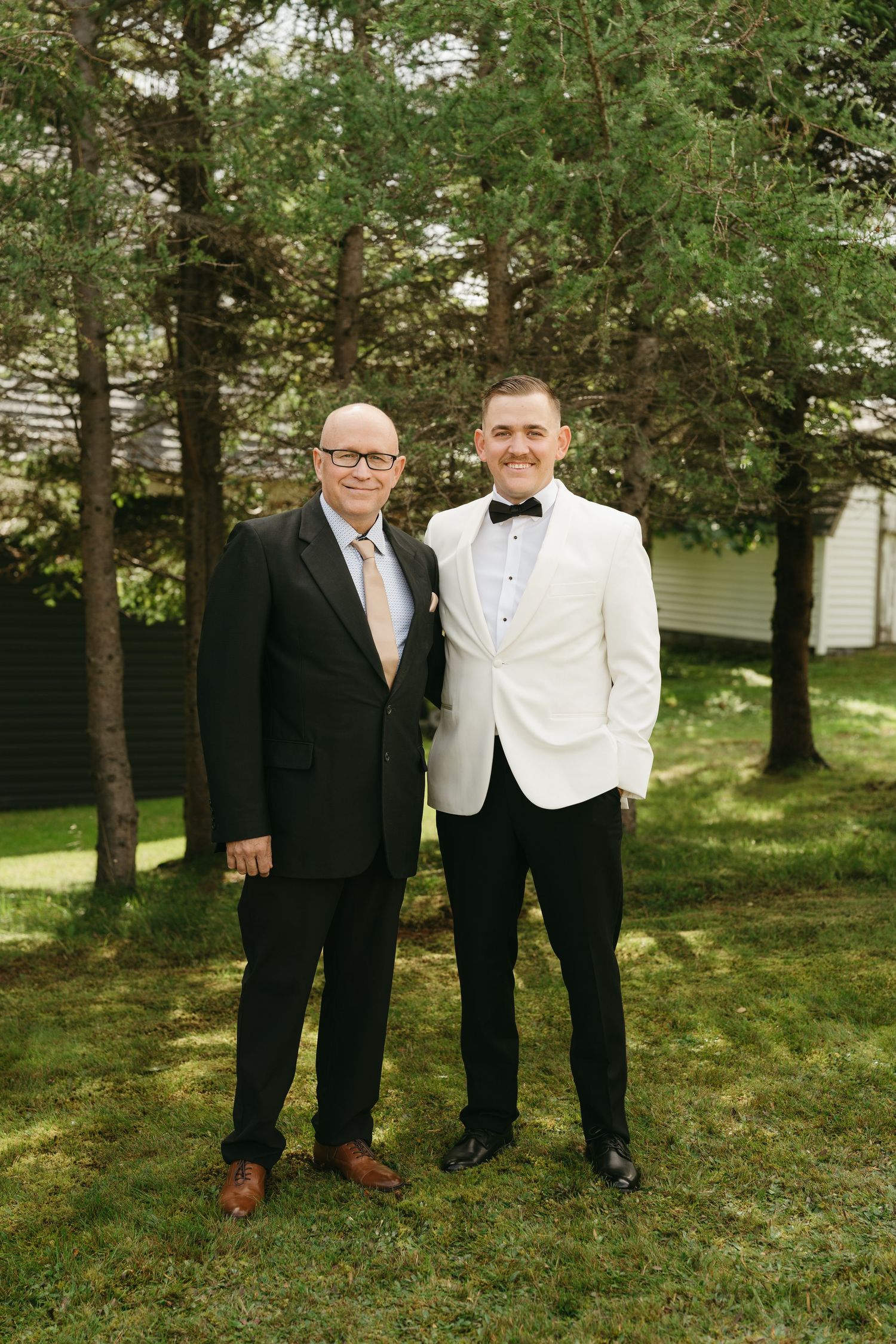Two people in formal attire pose together on a lawn with trees in the background.