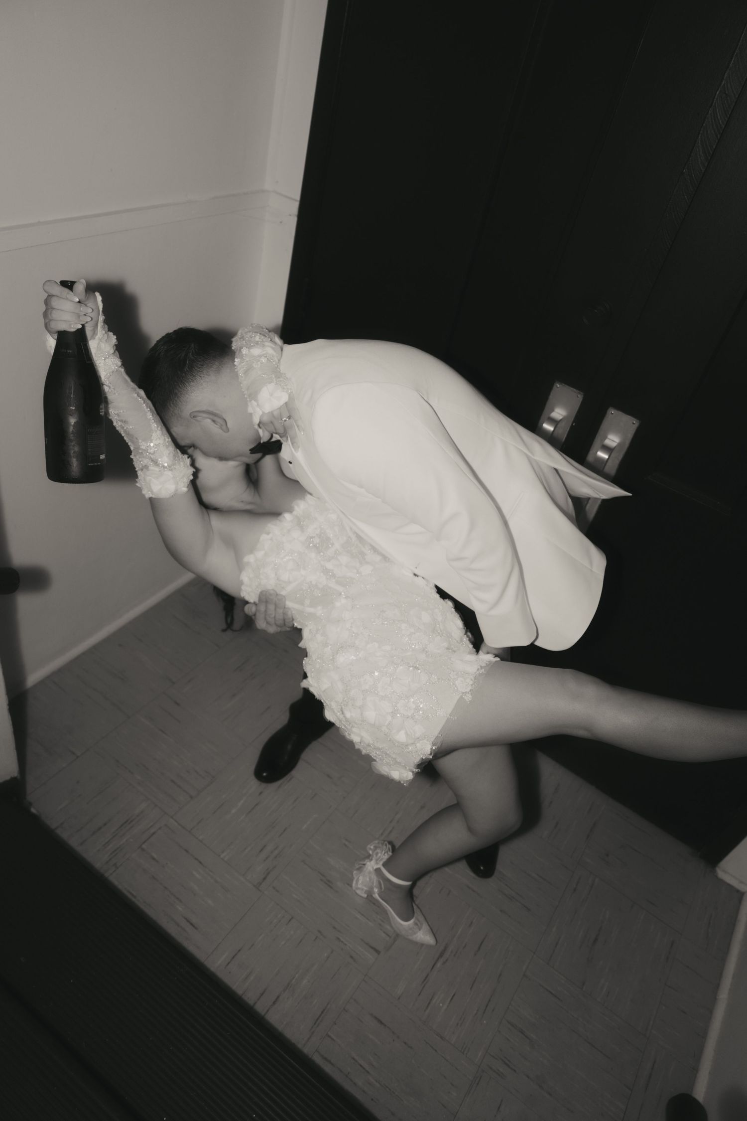 A couple in formal wedding attire share a romantic dip pose while holding champagne at an indoor celebration.