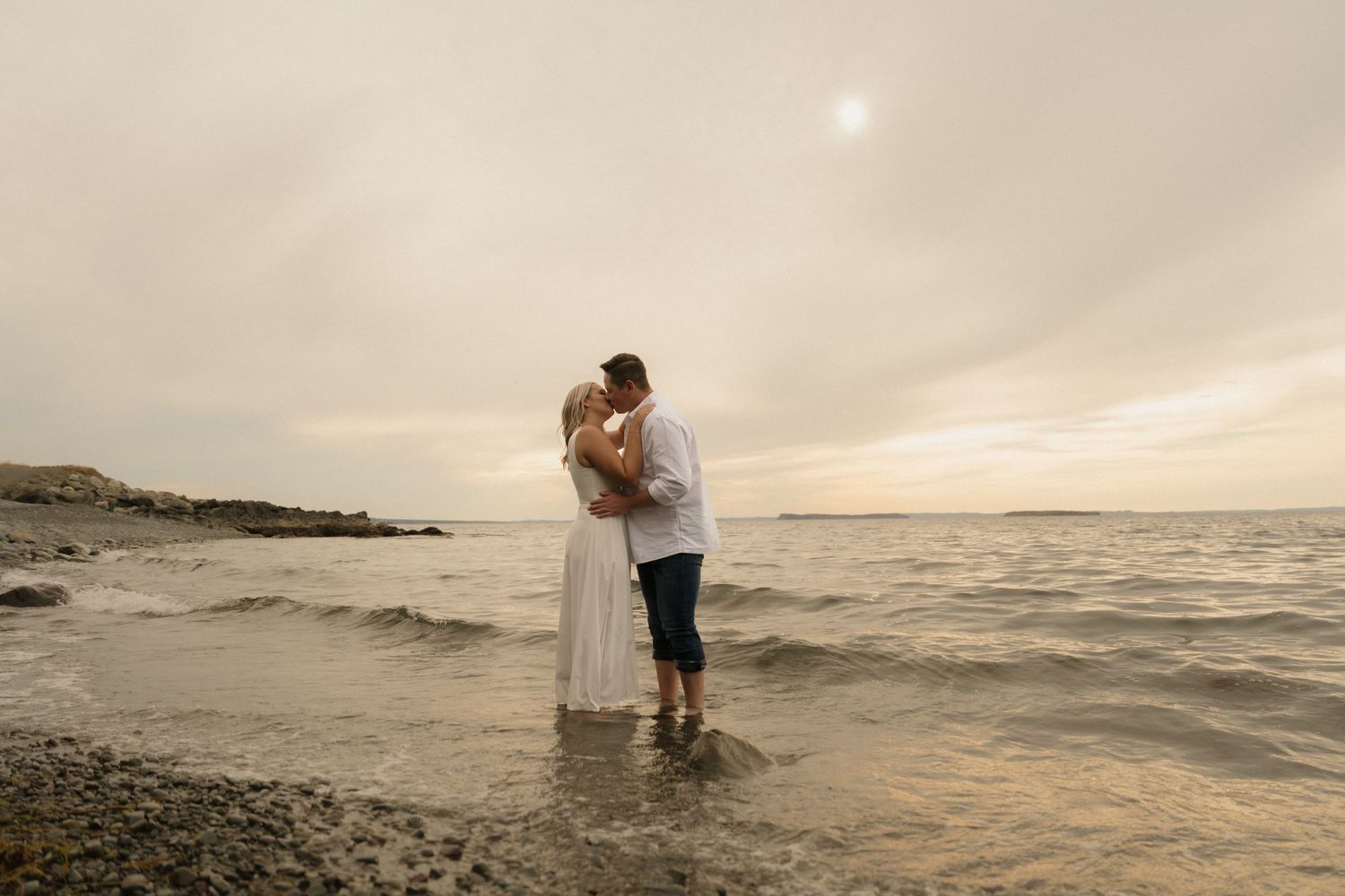 A couple embraces on a rocky beach at sunset with misty skies and ocean waves in the background.