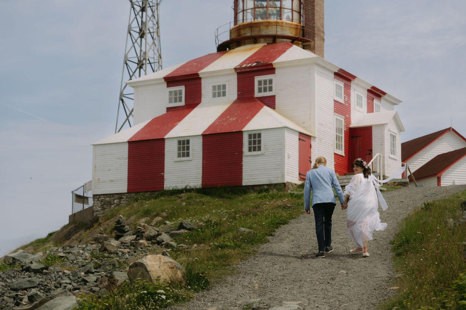 Two people walk up a path towards a red and white lighthouse on a rugged coastal cliff under cloudy skies.