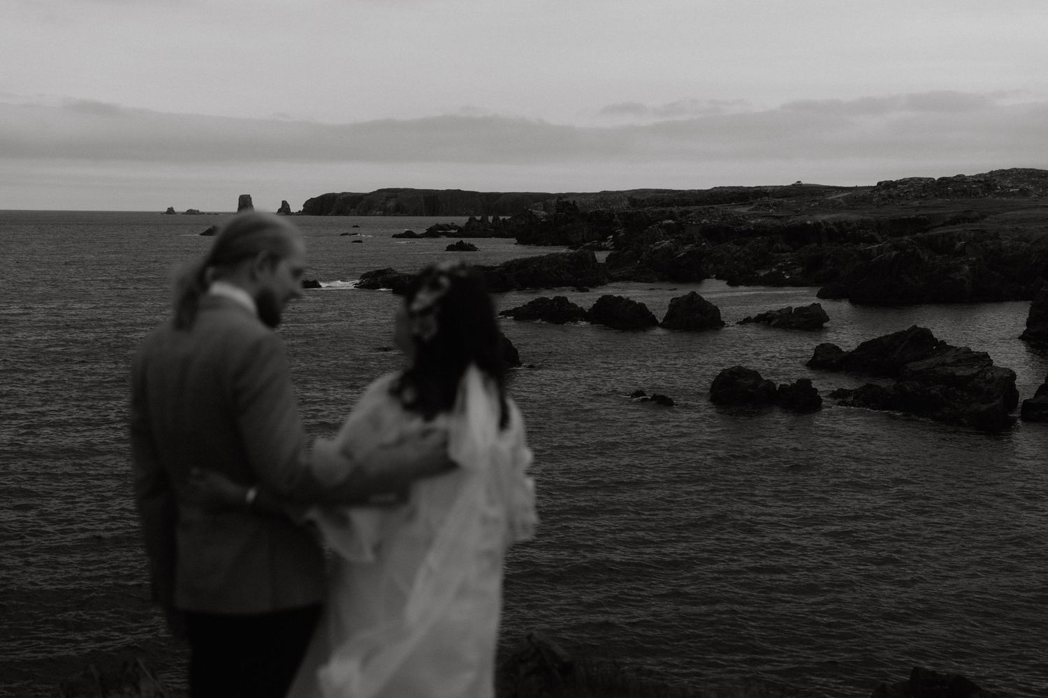 A black and white coastal scene with two figures standing on rocky cliffs overlooking the ocean at dusk.