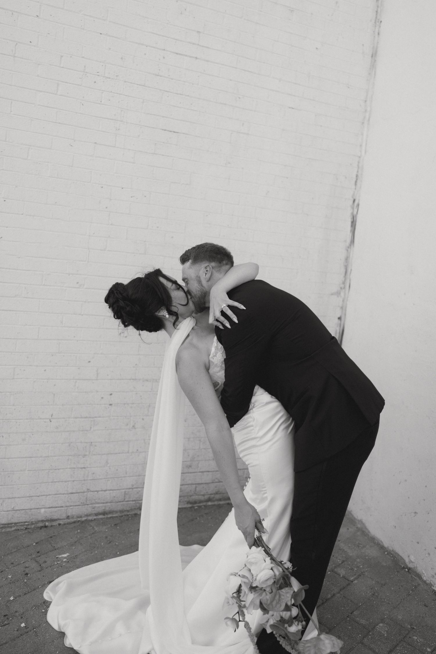 Black and white photo of a bride and groom sharing a romantic dip kiss against a white brick wall.