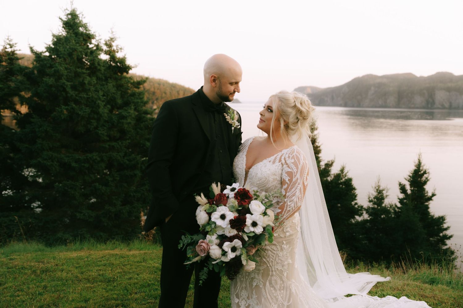 Newlyweds pose against a scenic coastal landscape with evergreen trees and water views.