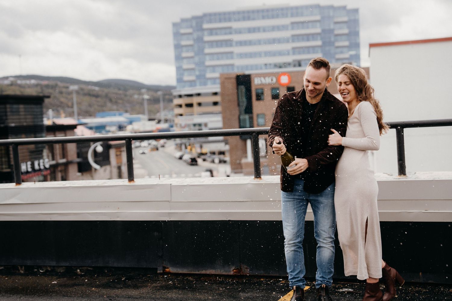 A couple embraces while standing on a rooftop deck with urban architecture visible behind them.