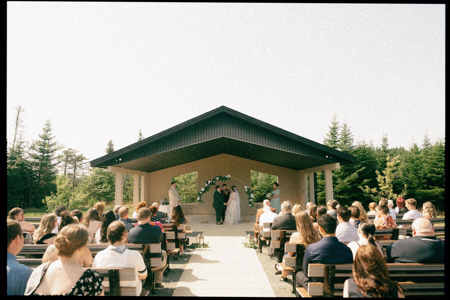 Outdoor wedding ceremony at a rustic venue with a covered gazebo and guests seated on wooden benches in a peaceful forest setting.