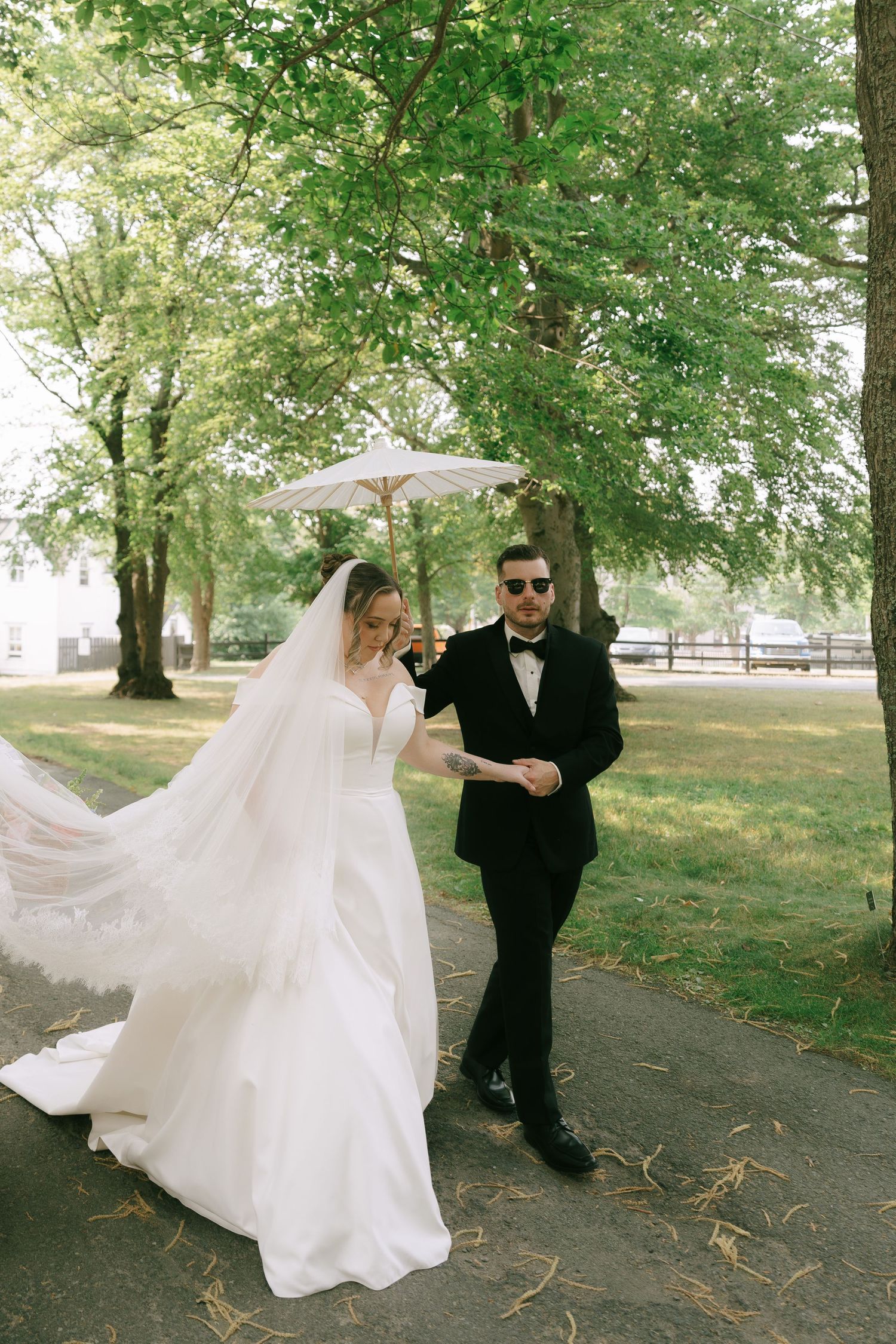 Bride in flowing white dress twirling outdoors under green trees.
