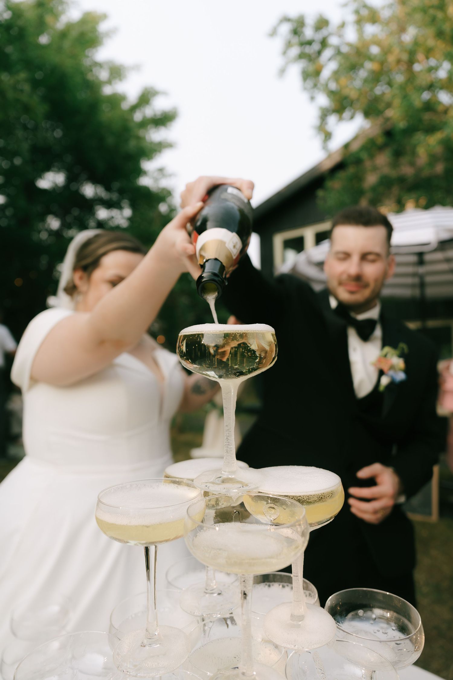 Pouring celebration champagne into a tower of coupe glasses at an outdoor wedding reception.