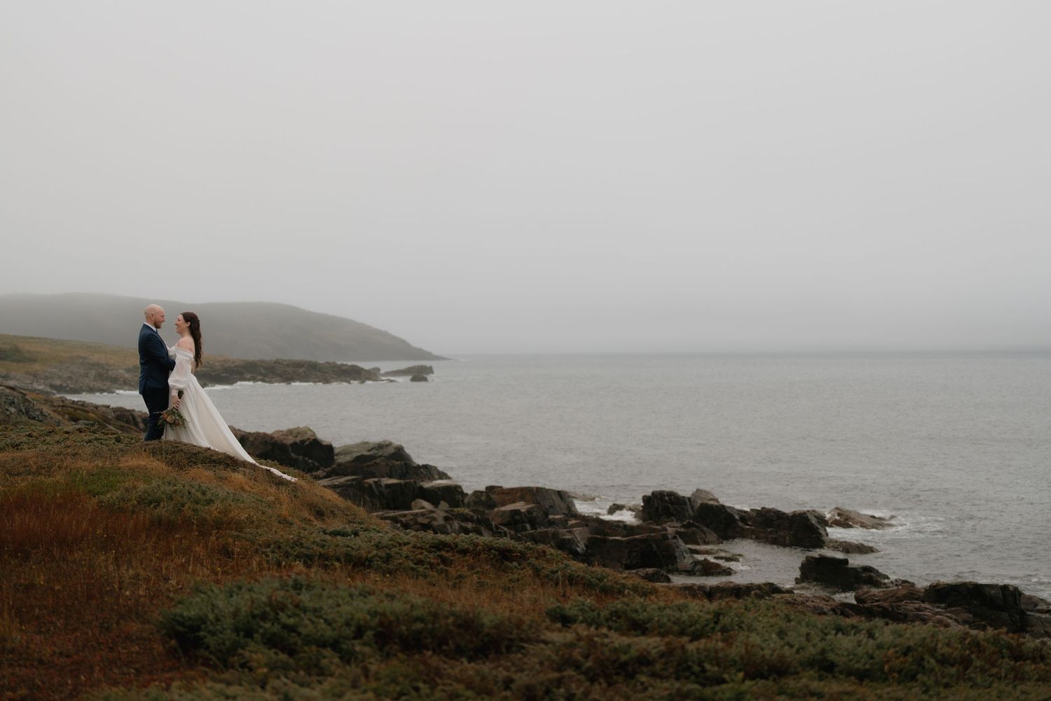 A couple in wedding attire stands on a misty coastal cliff overlooking the ocean on their special day.