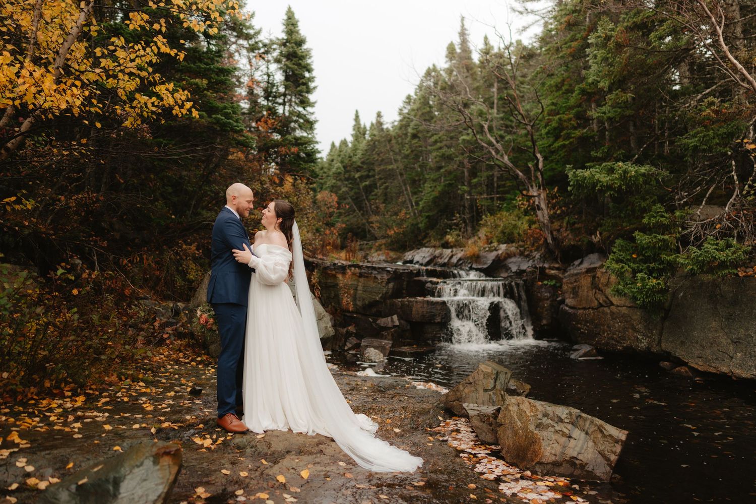 A couple in formal attire embraces near a scenic waterfall surrounded by vibrant autumn foliage in a forest setting.