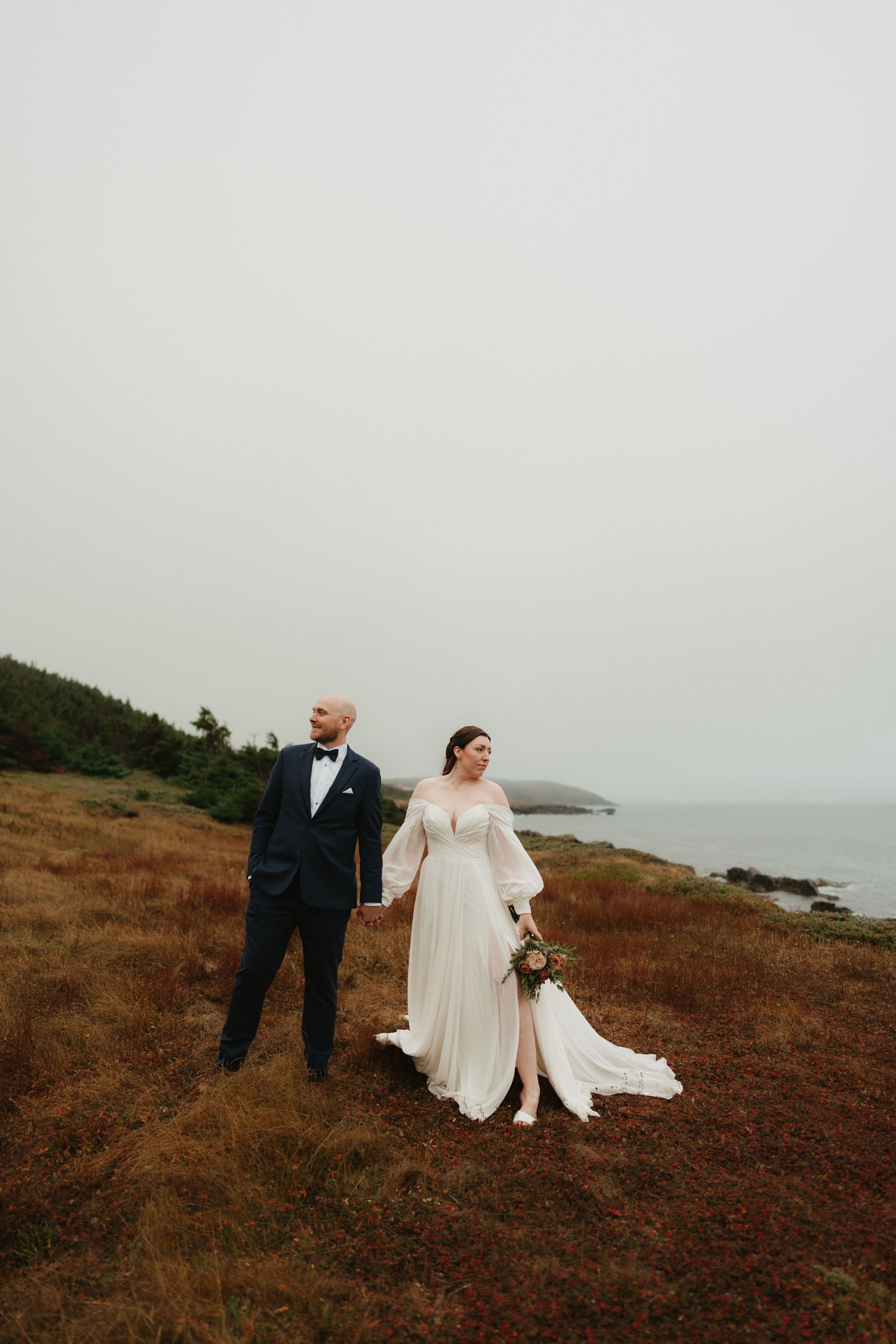 Wedding couple embracing on rugged oceanside terrain with dramatic cloudy sky backdrop.