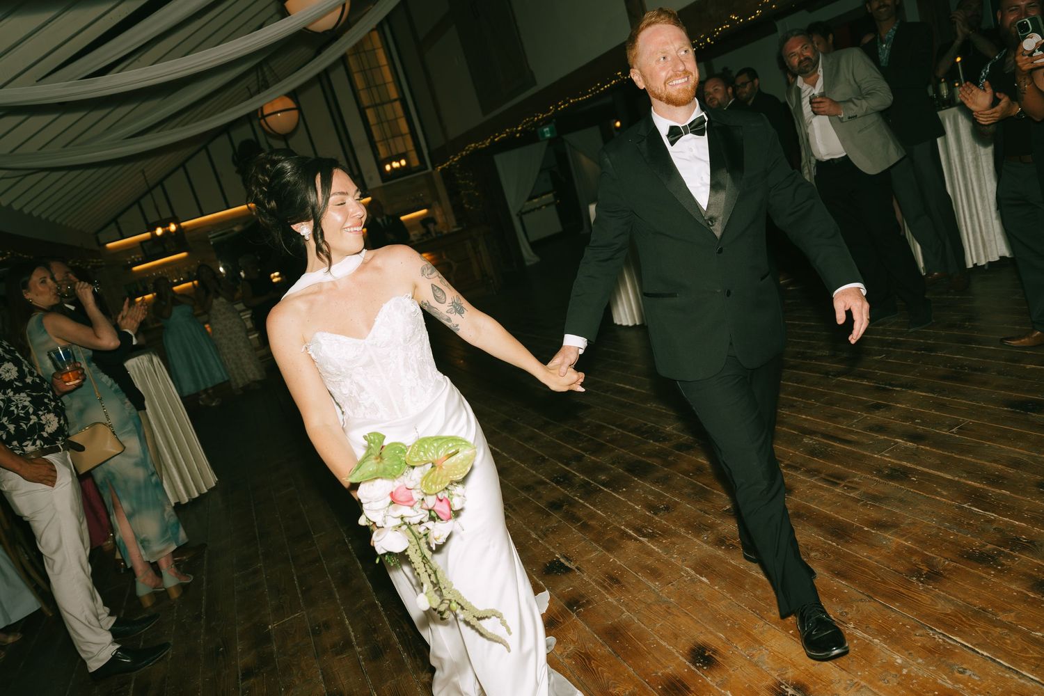 Bride and groom holding hands walking onto dance floor at wedding reception, bride holding pink bouquet.