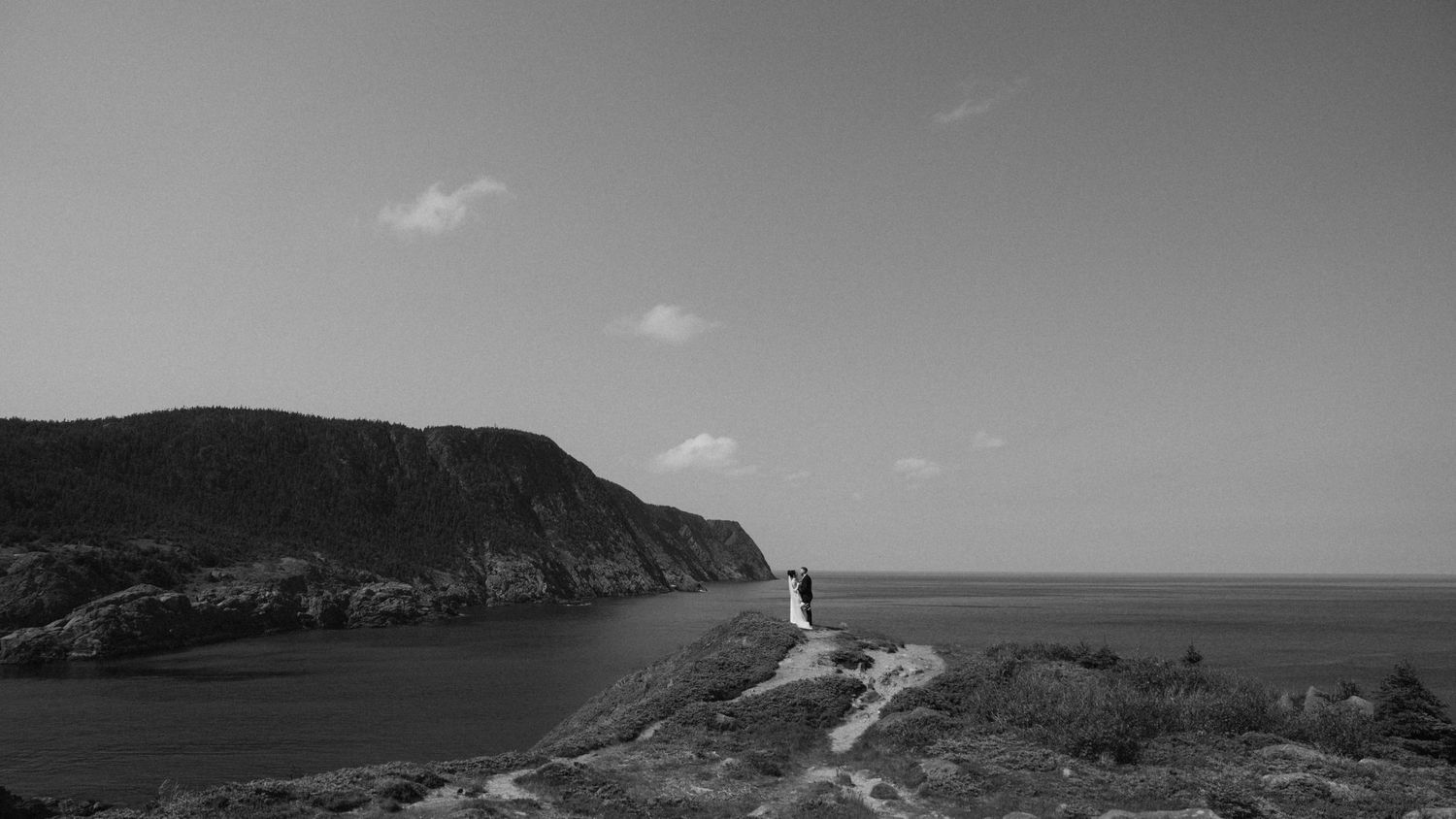 Black and white photo of a person standing on coastal cliffs overlooking a serene ocean bay with dramatic hillside backdrop.