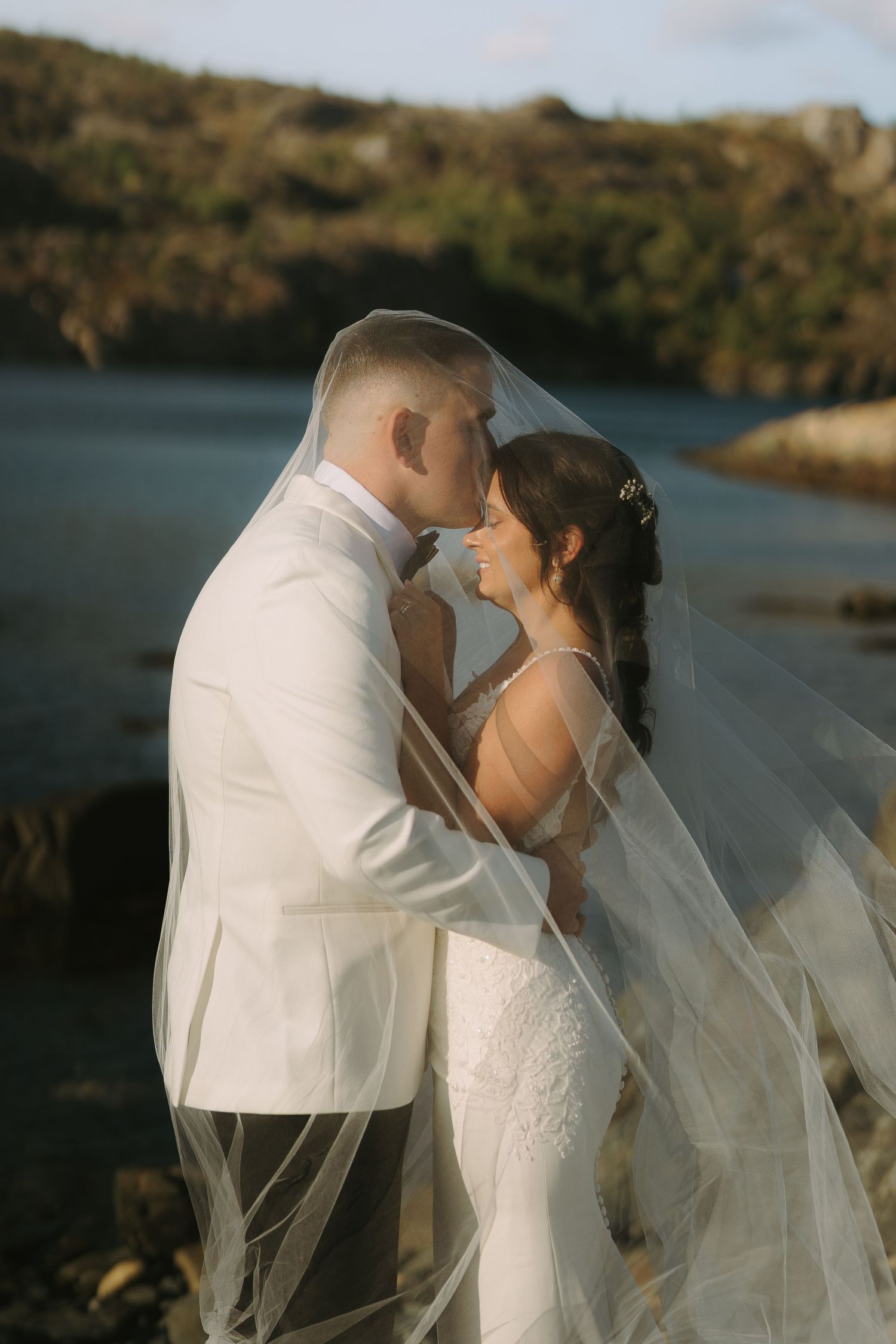 A romantic wedding couple shares intimate moments by a scenic waterfront during golden hour sunset.
