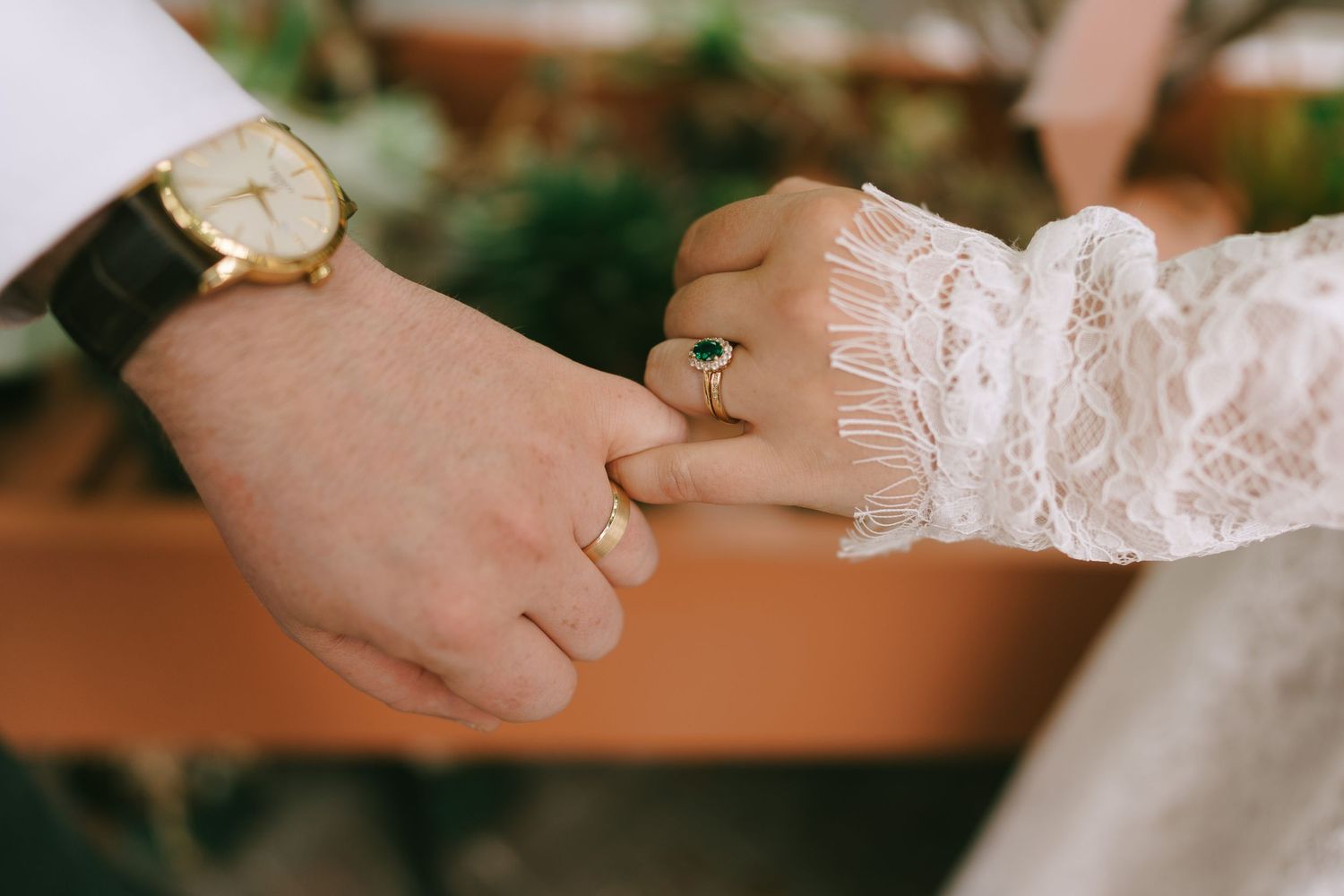 Bride and groom holding hands, showing gold wedding rings and a green gemstone ring, with a gold watch on the groom.