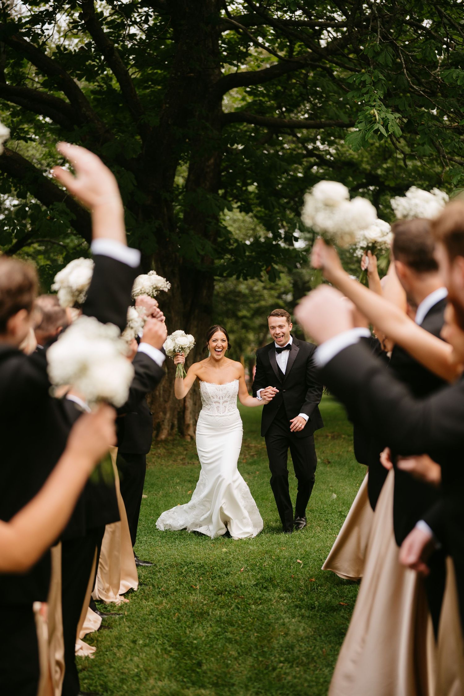 Bride and groom walk joyfully through an aisle of wedding party holding white bouquets aloft outdoors.