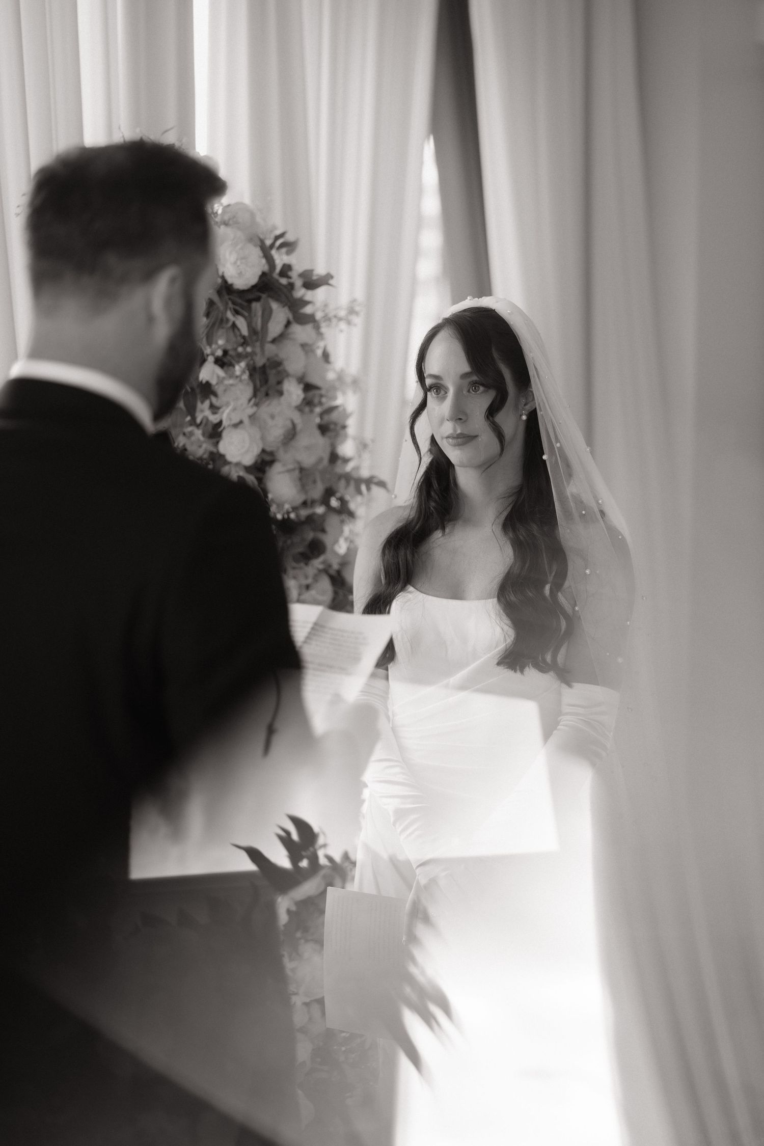 Black and white wedding photo of a bride in a white gown and veil reading vows to her groom during ceremony.