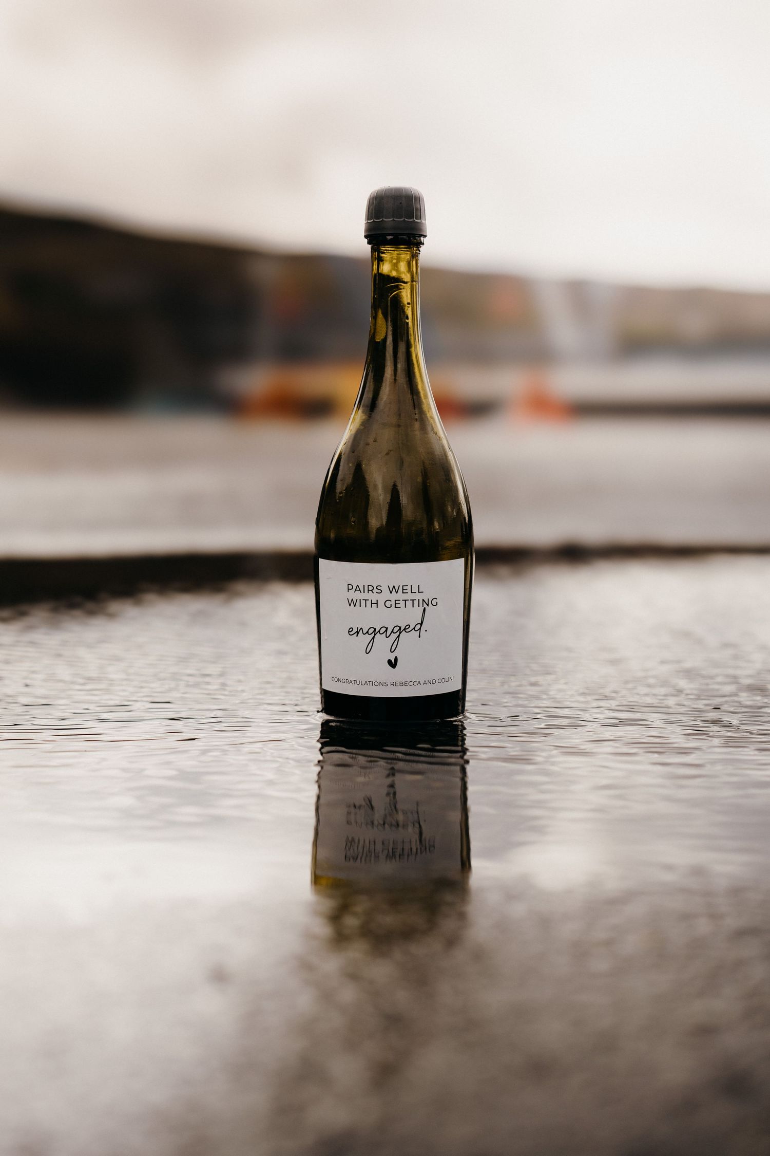 Wine bottle with engagement announcement label reflecting in puddle of water.