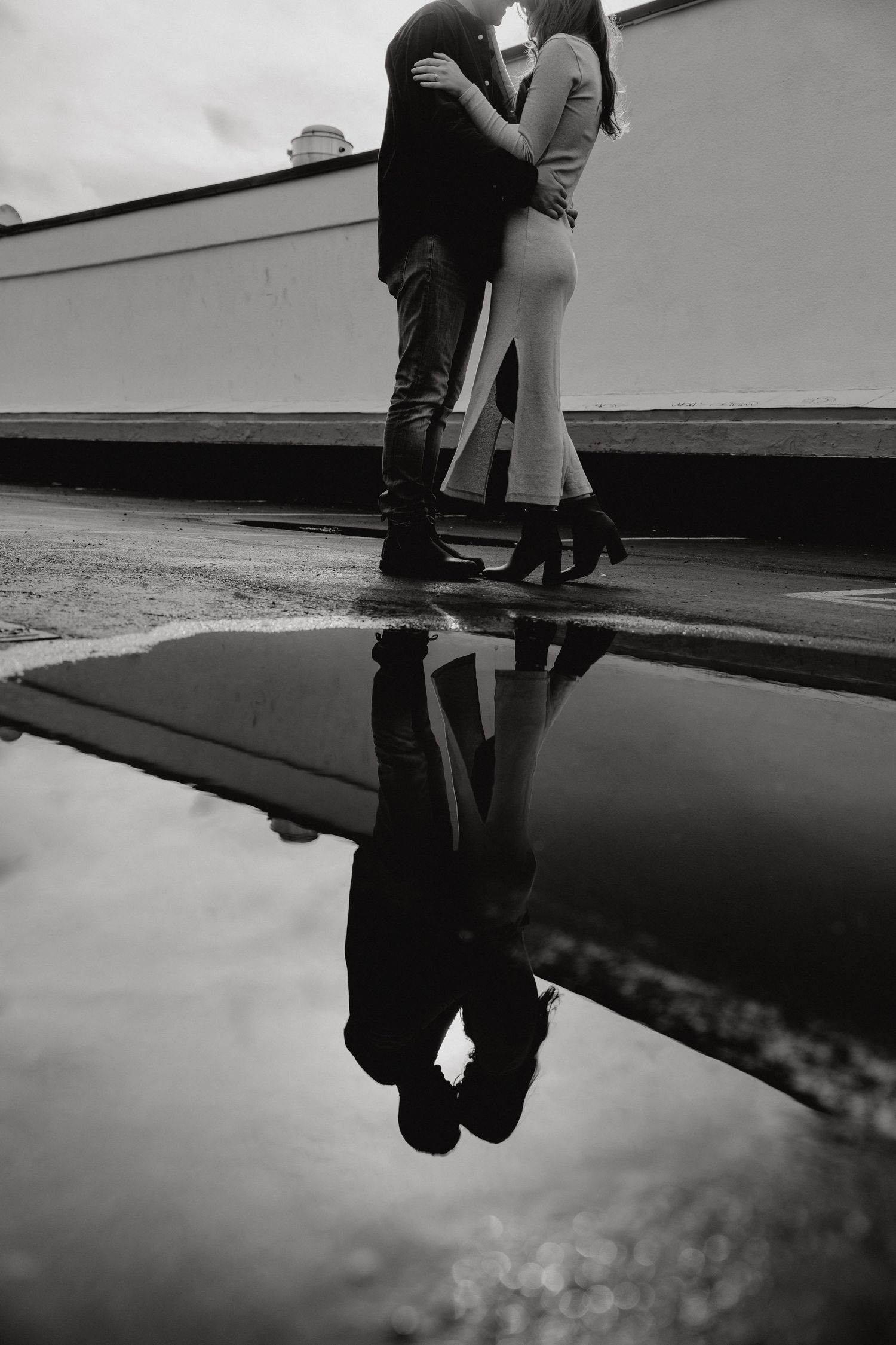 Moody black and white reflection of a couple's embrace in urban puddle.