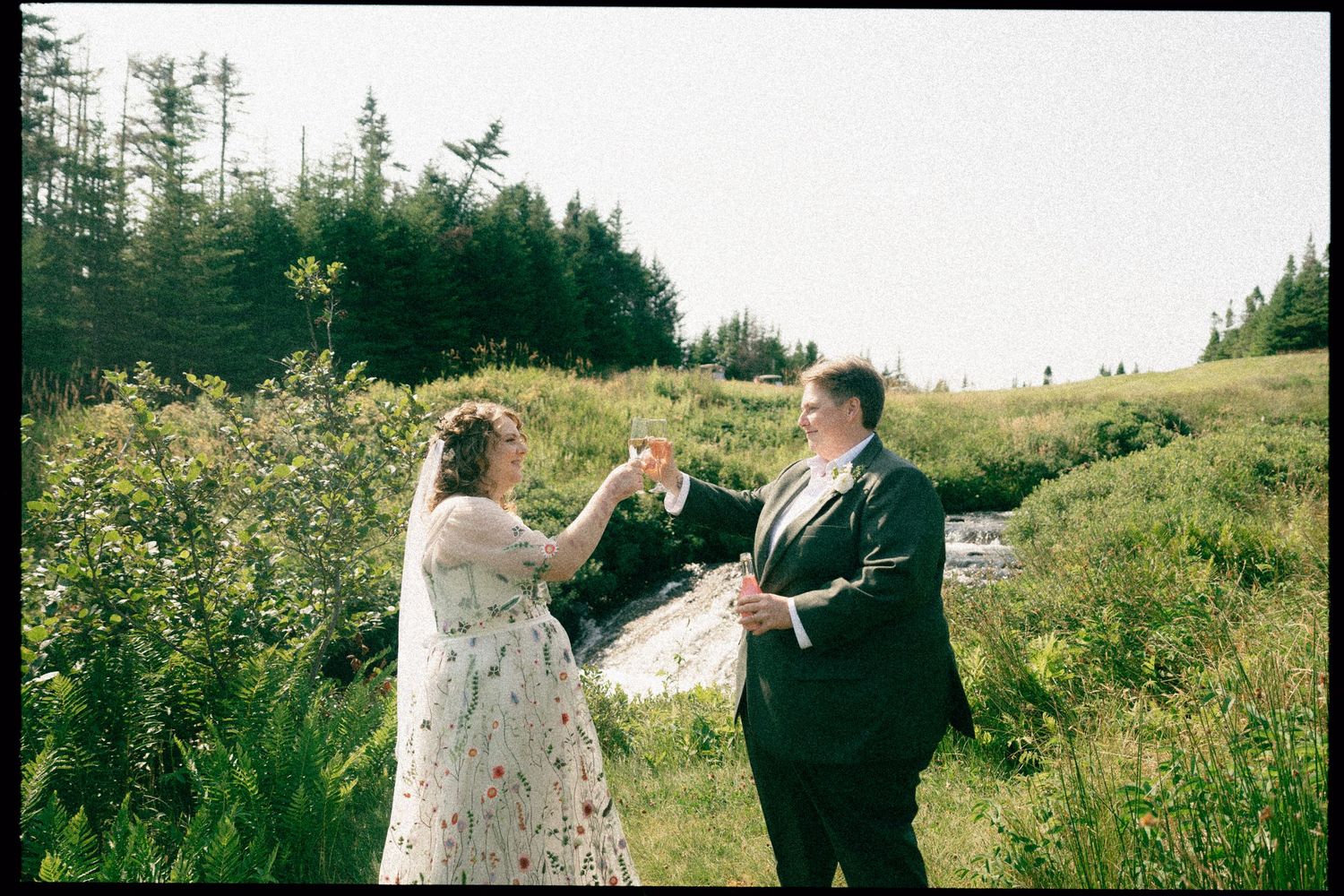 A couple in formal attire celebrates outdoors in a scenic field with evergreen trees and greenery.