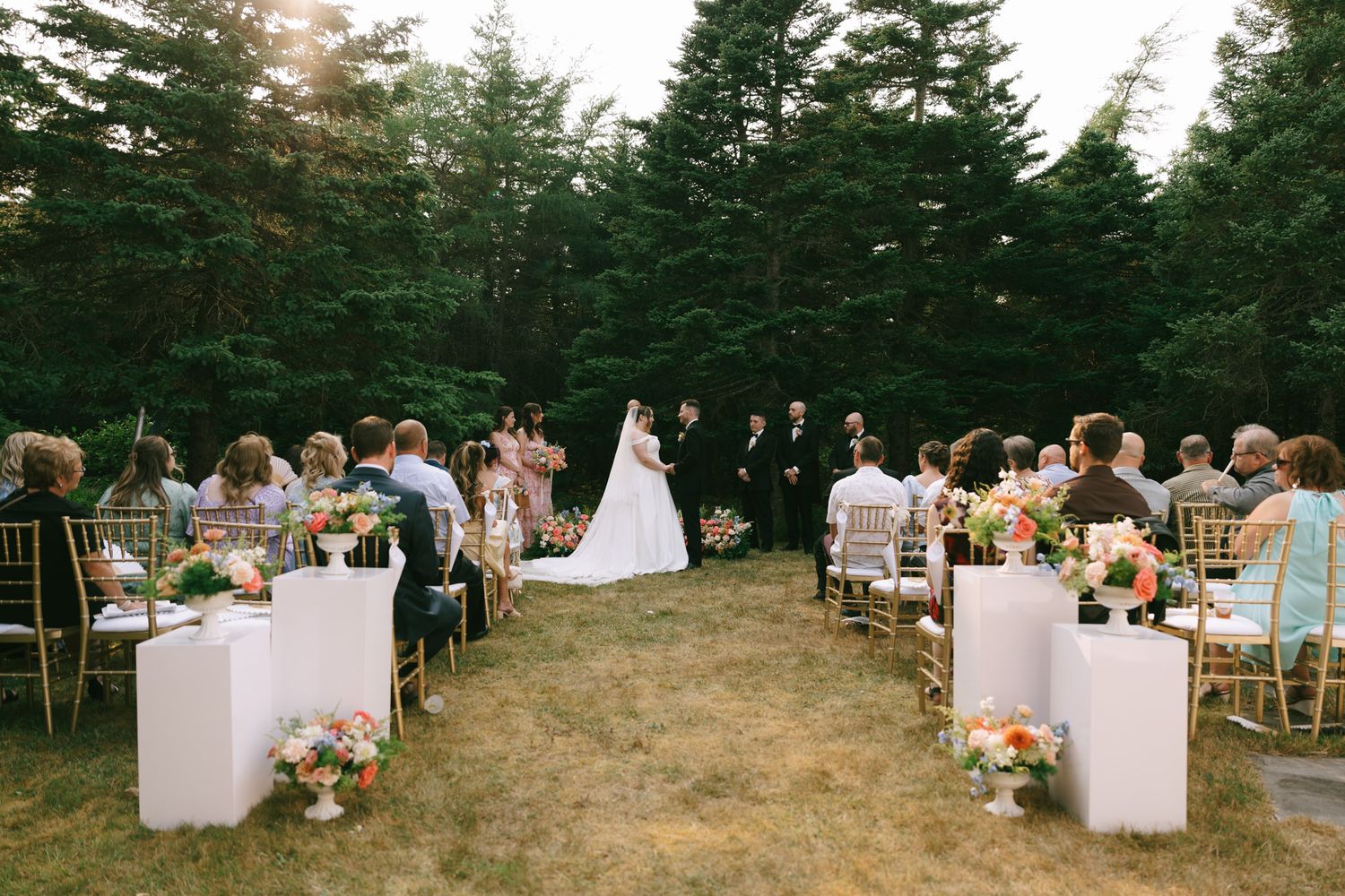 Panoramic view of an outdoor wedding reception setup in a pine forest with white table settings.