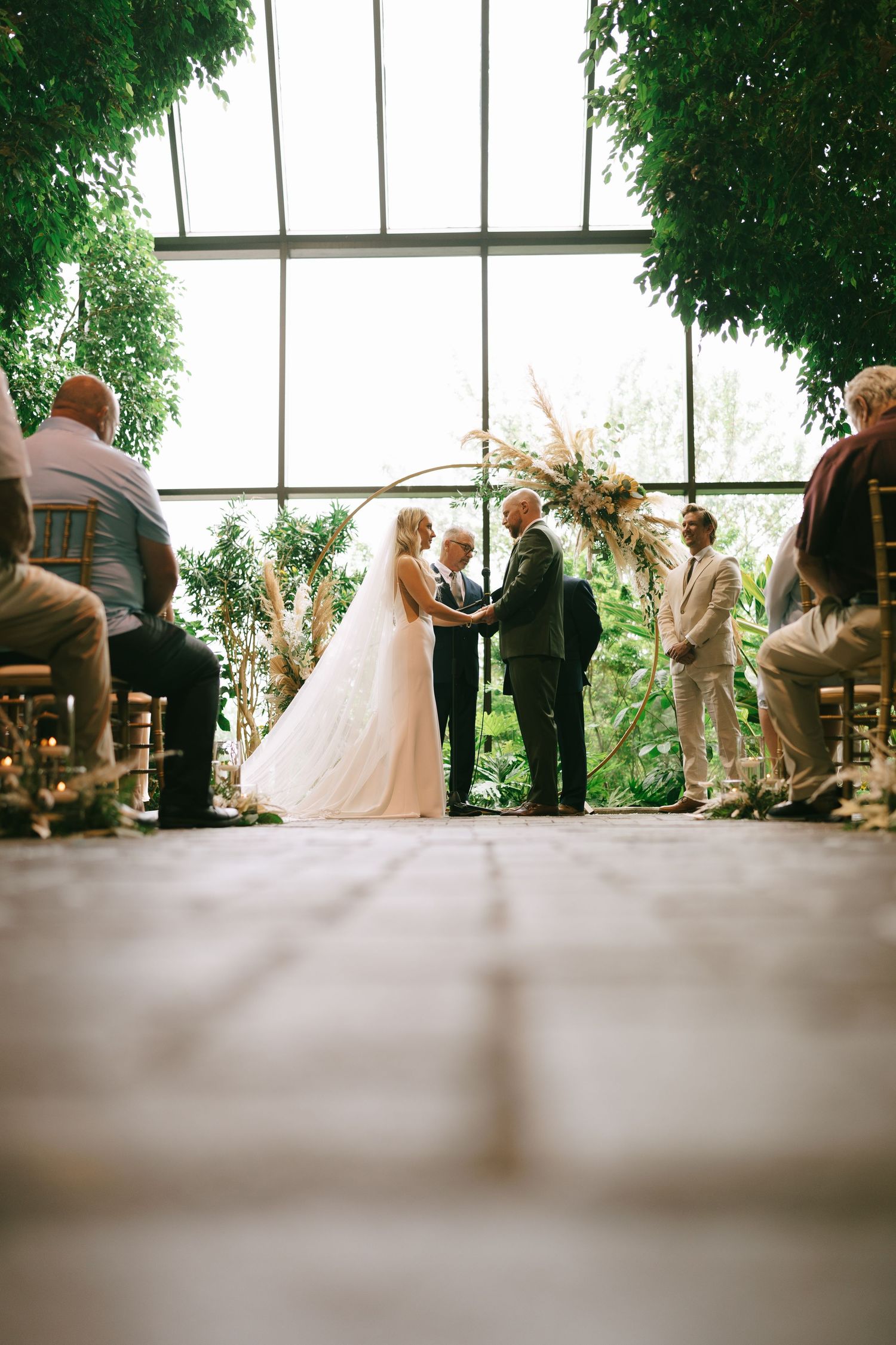 A wedding ceremony in progress inside a bright venue with large windows and green foliage.