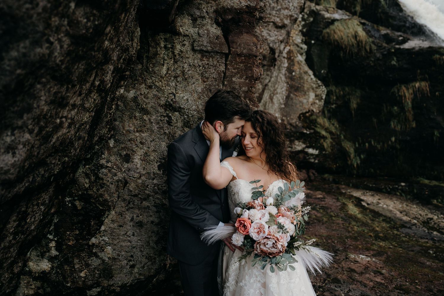 Couple poses against rugged rock formation with cascading waterfall in background.