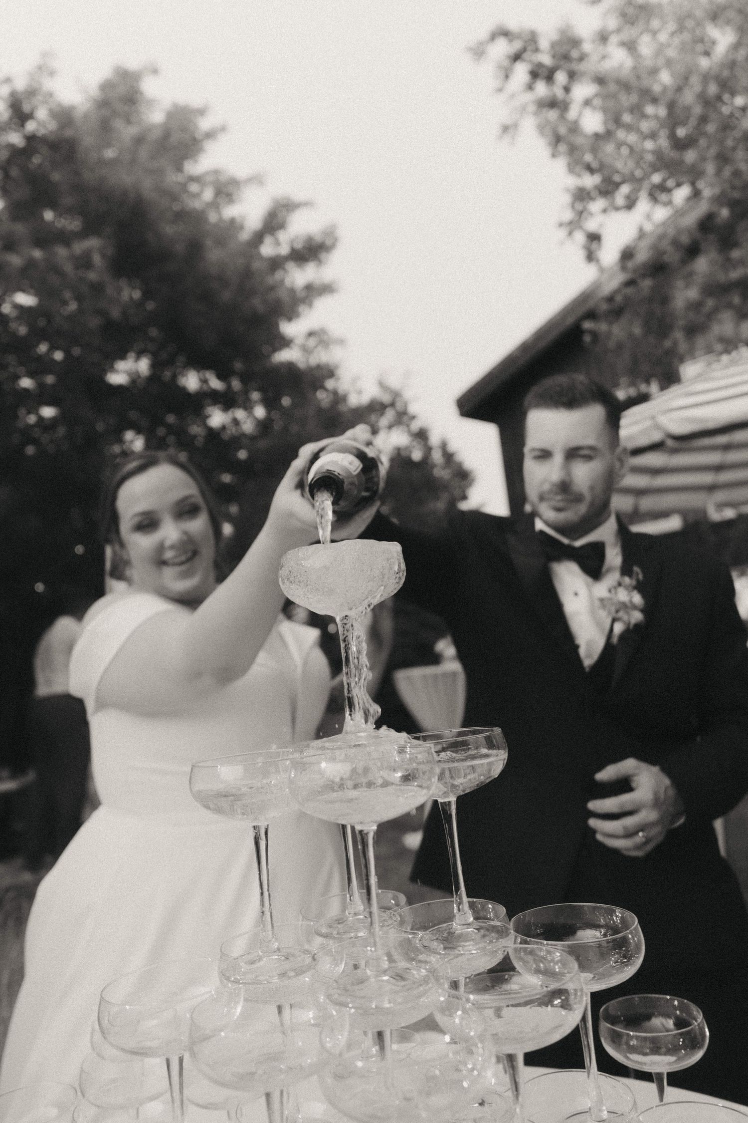 Elegant champagne tower being filled at an outdoor wedding celebration with trees in background.