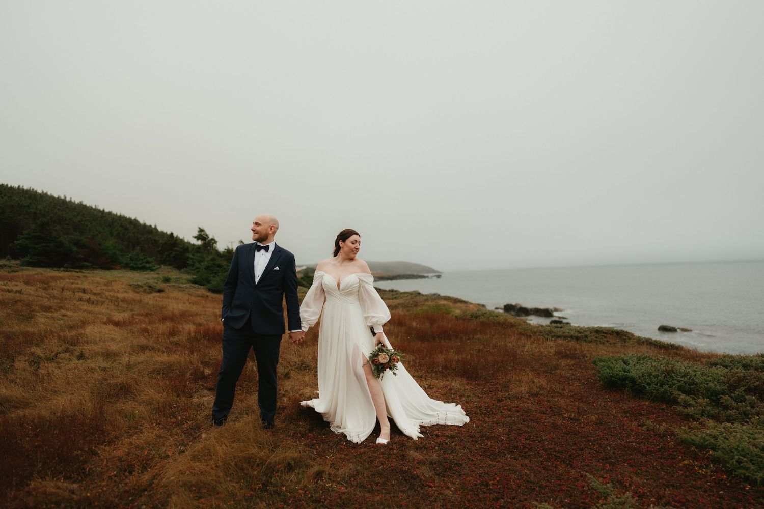 Romantic coastal wedding portrait with couple standing on rustic brown hillside by the sea.