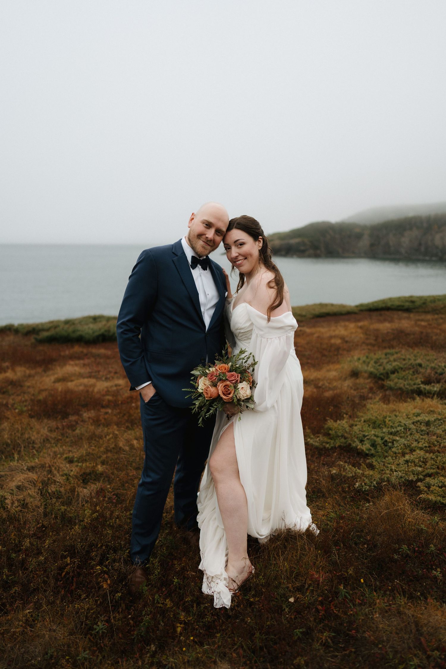 A couple in wedding attire embrace on a misty coastal cliff with autumn-colored grass and a moody ocean backdrop.