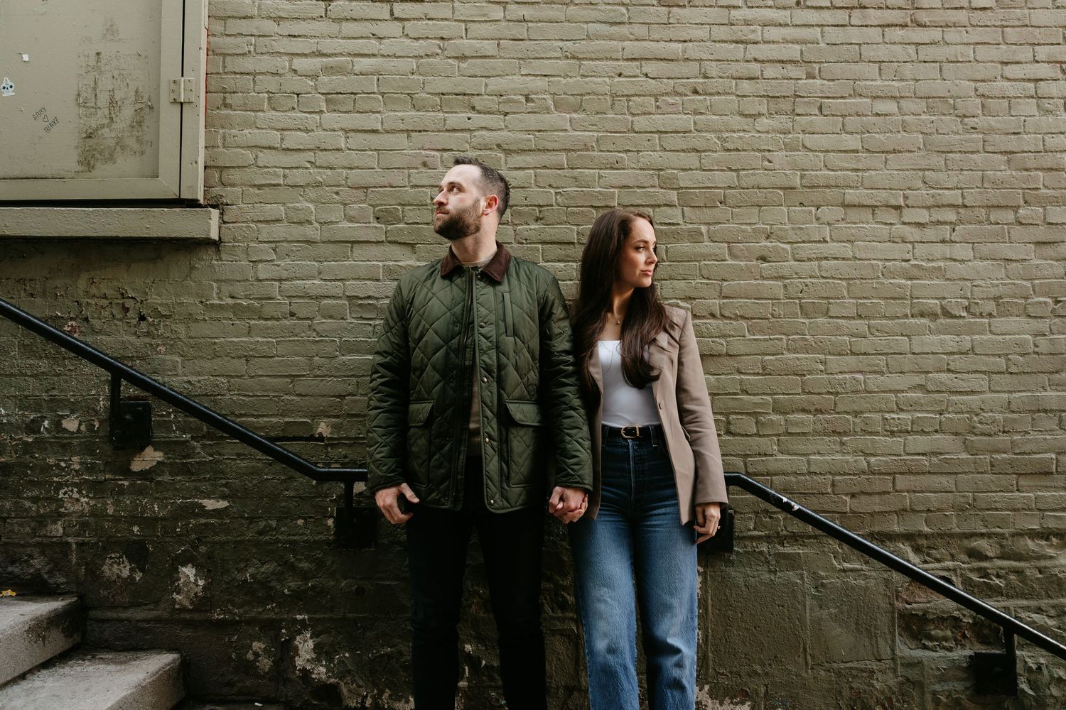 Couple standing back-to-back holding hands against a brick wall near outdoor stairs in urban setting.