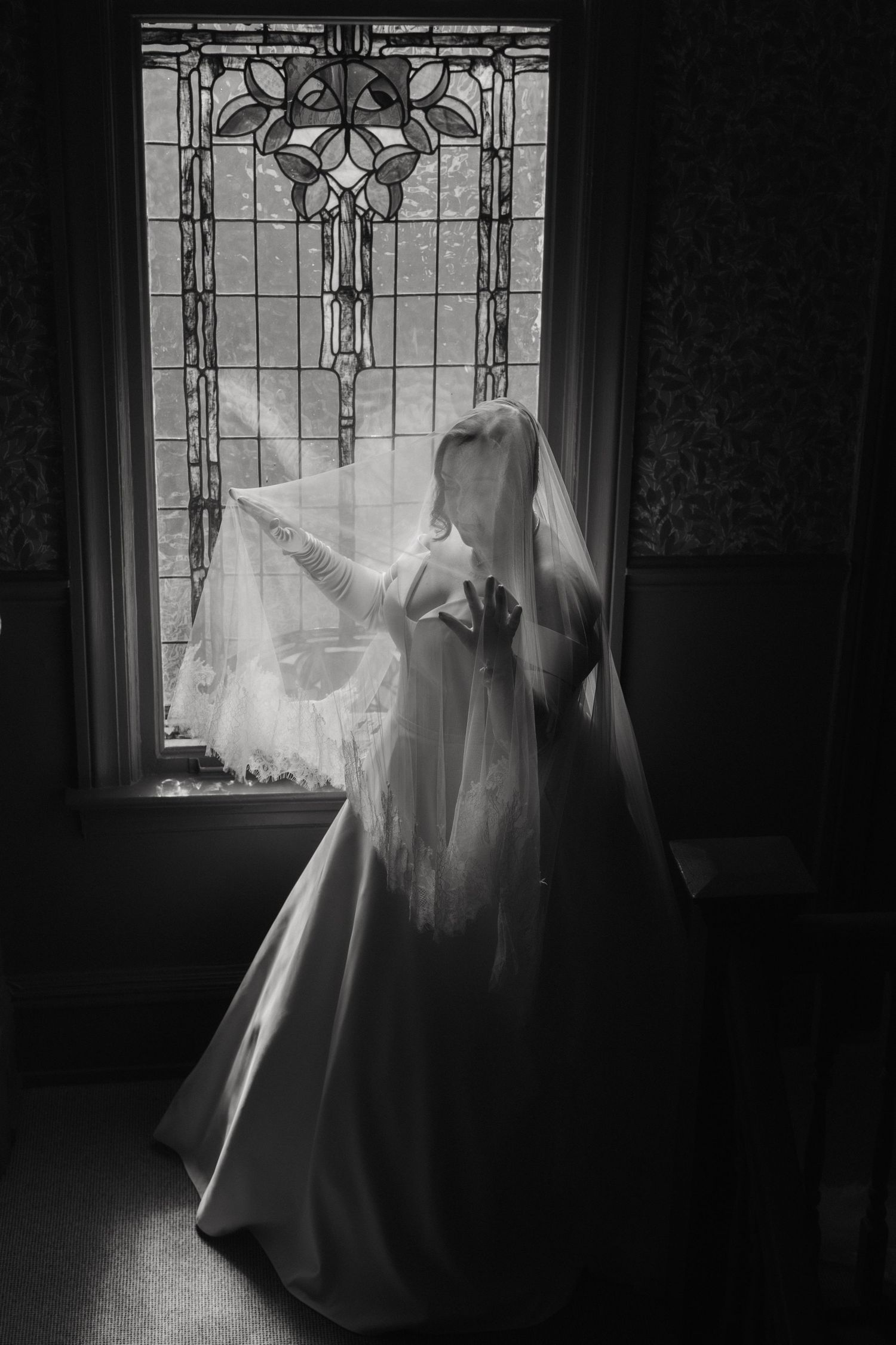 A flowing white wedding dress silhouetted against a stained glass window in dramatic black and white.