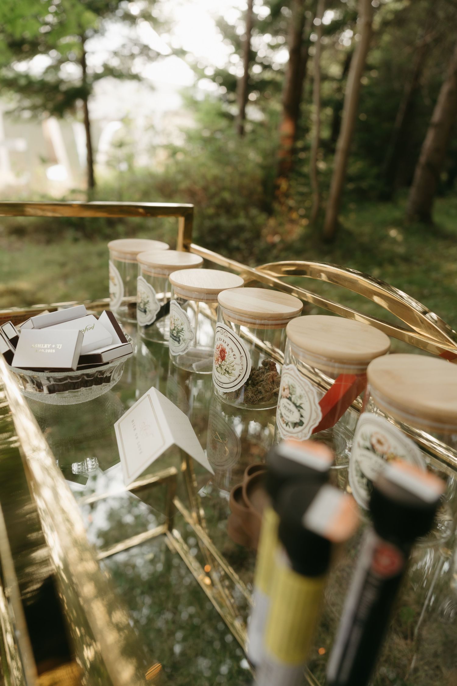 Row of wine bottles and glasses arranged in outdoor woodland setting.