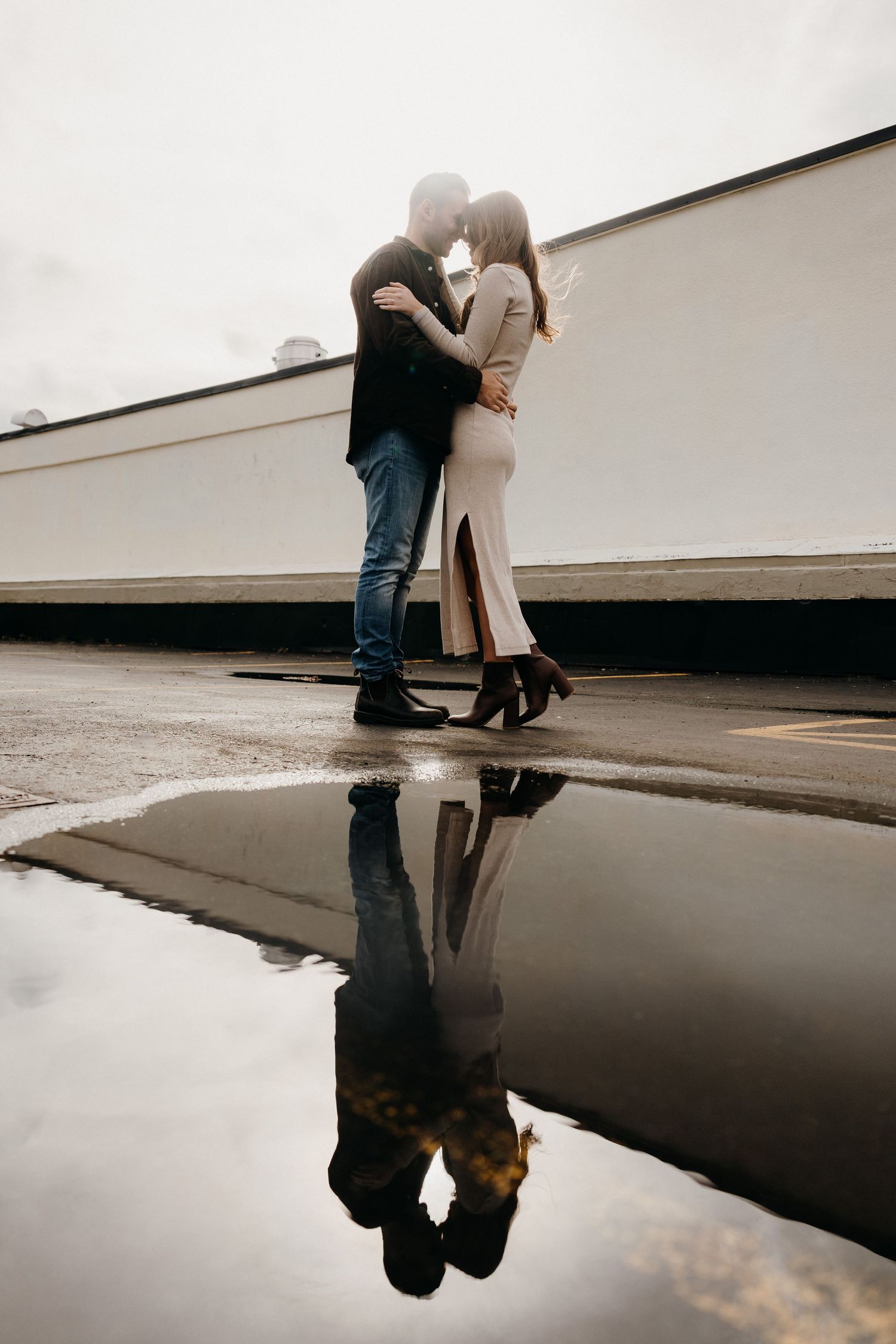 A couple's reflection appears in a puddle while embracing near white industrial walls.