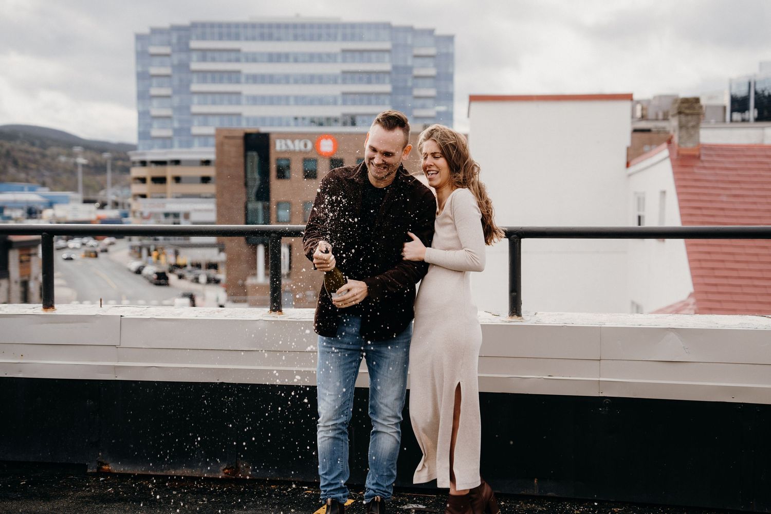 Two people stand together on a rooftop overlooking buildings on a cloudy day.
