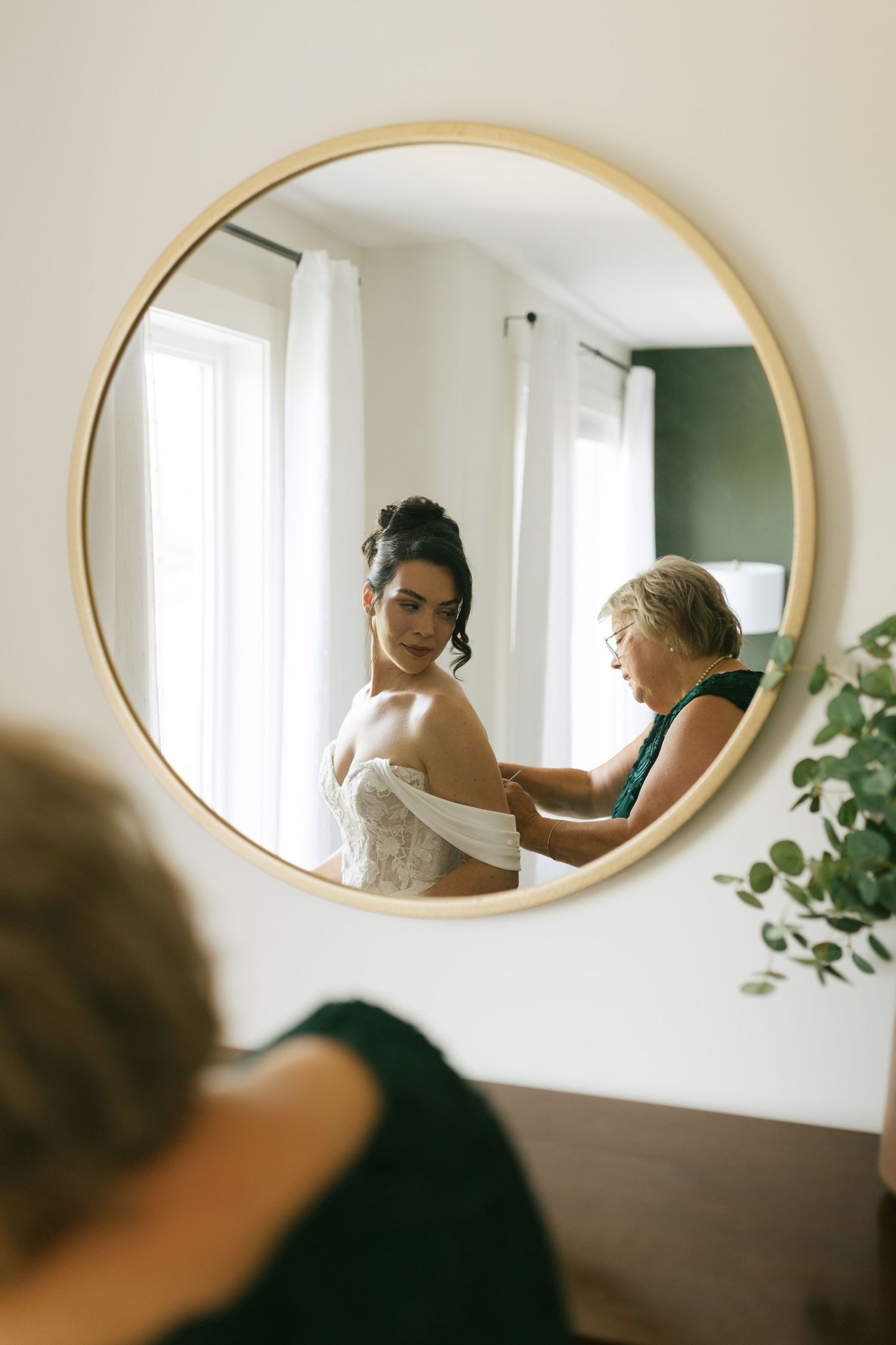 Bride getting help with her wedding dress in a round gold-framed mirror reflection during bridal preparations.