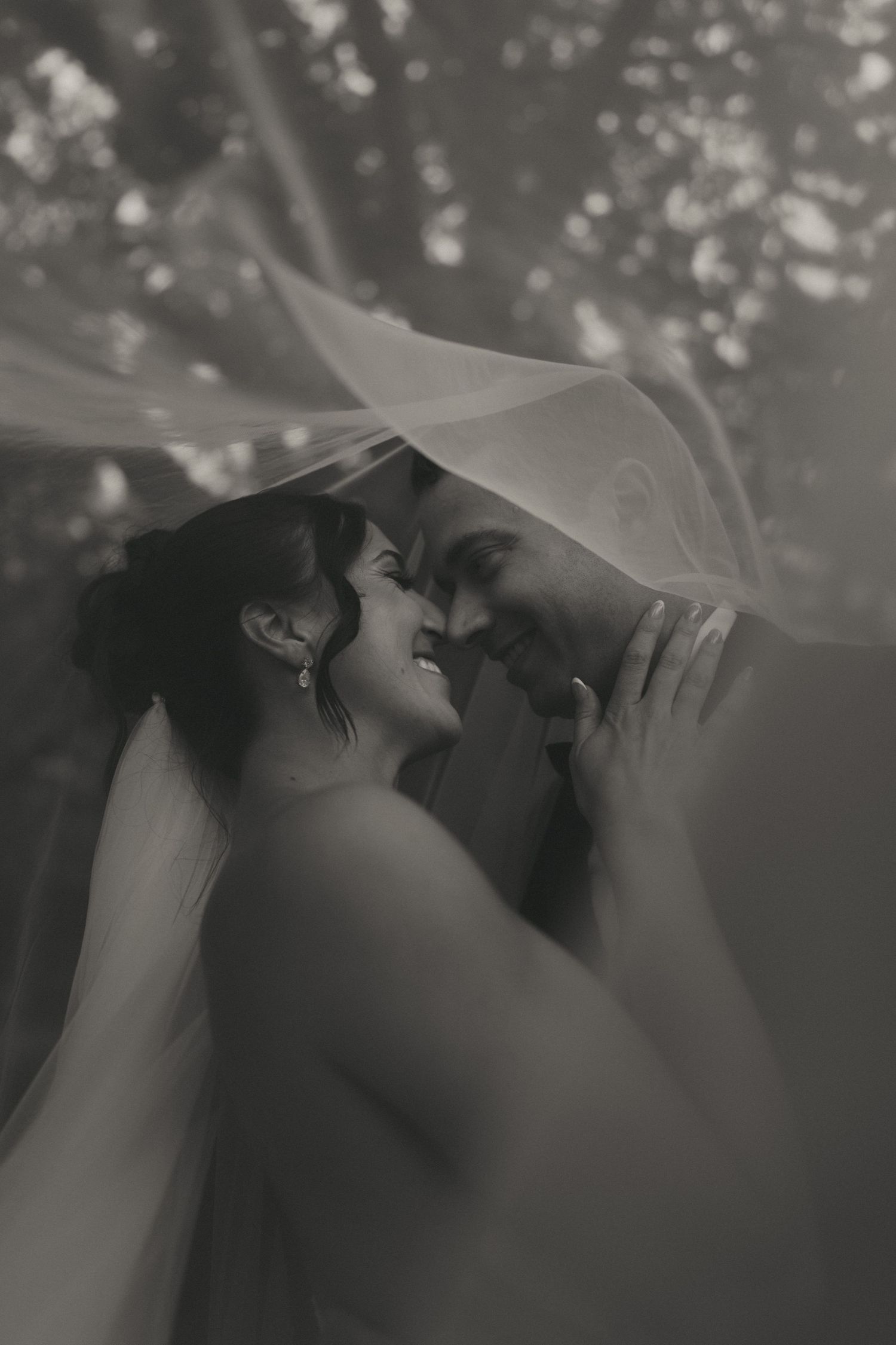 Black and white photo of a bride and groom sharing an intimate moment under a flowing veil outdoors.